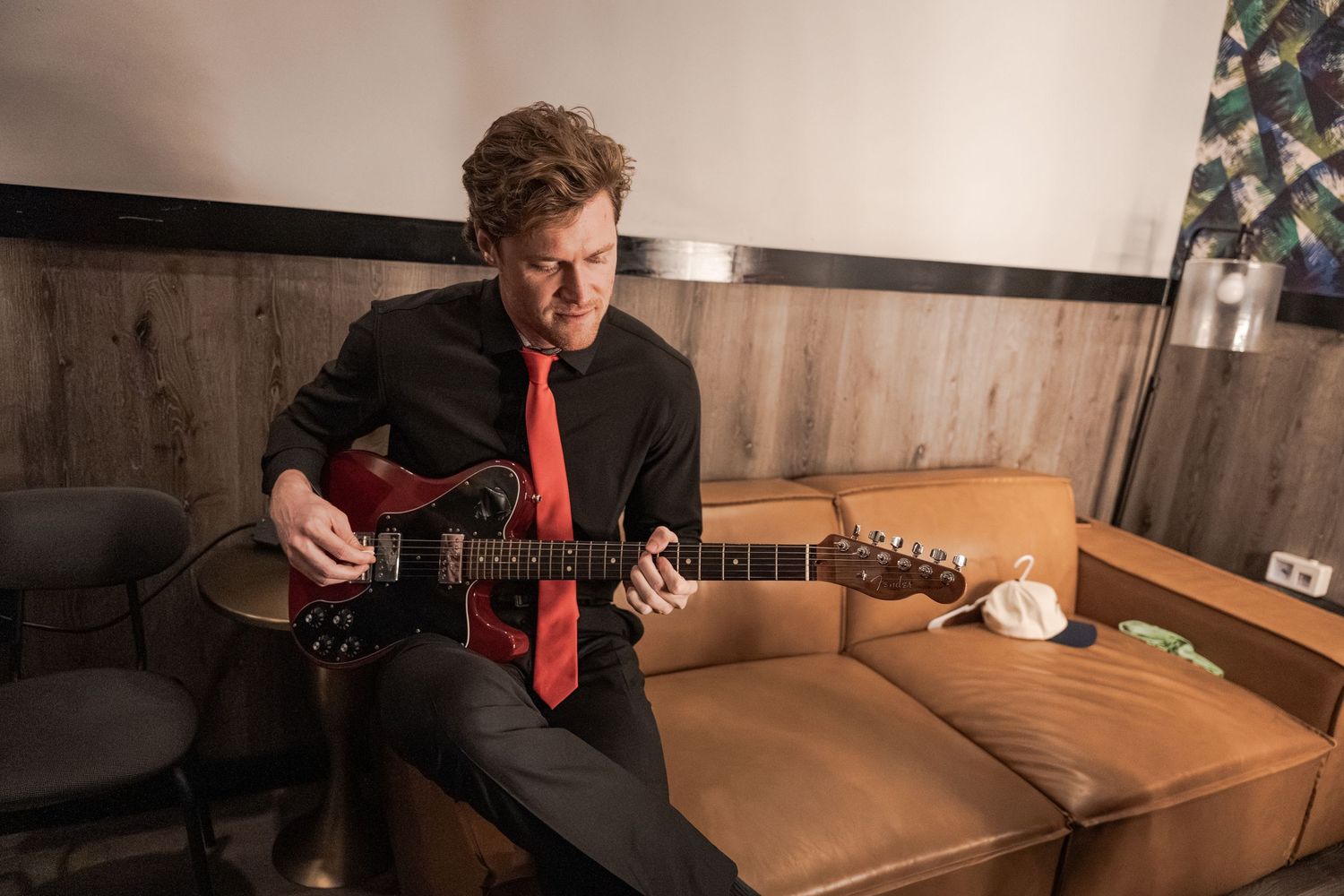 Man in black suit and red tie playing electric guitar on brown leather couch in modern room.