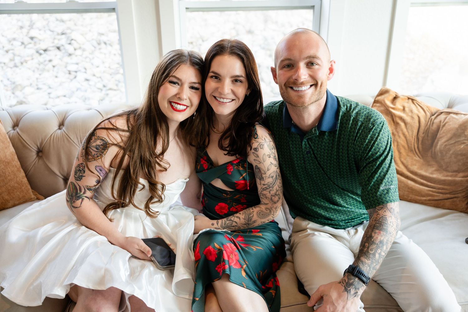 Three smiling people with tattoos sit together on a beige sofa in a bright, cozy living room setting.