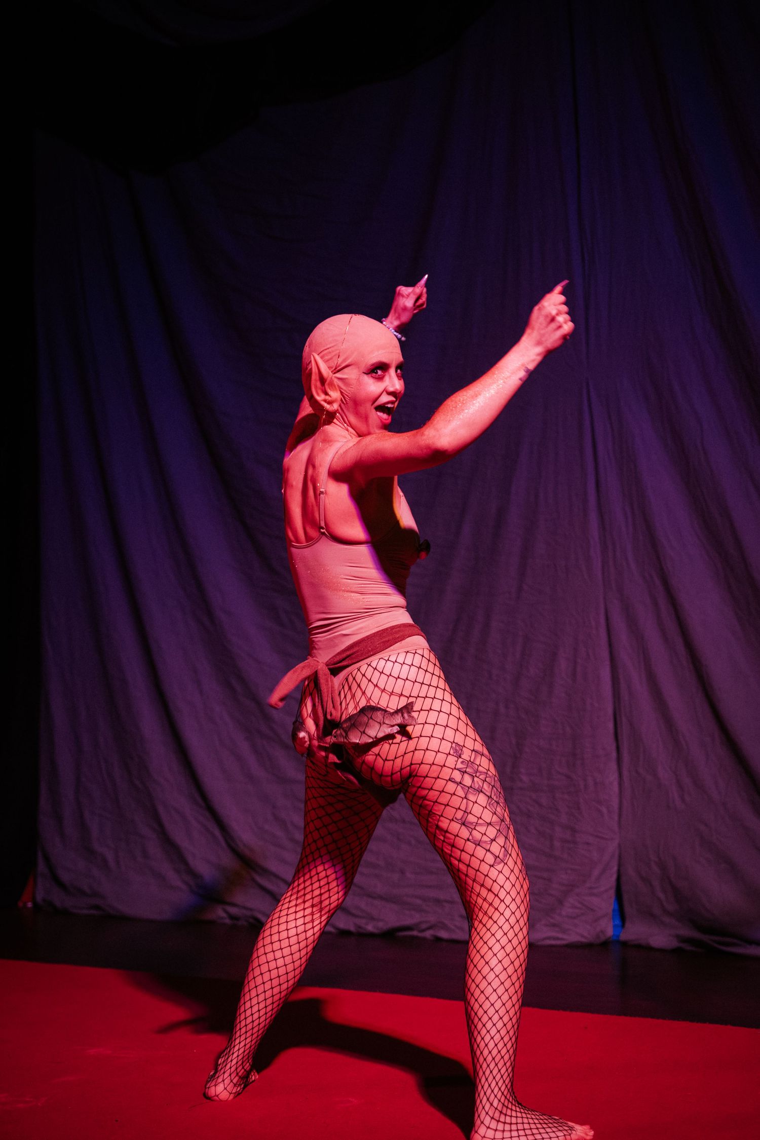 Performer in fishnet tights and body suit poses dramatically on red stage floor under pink lighting.