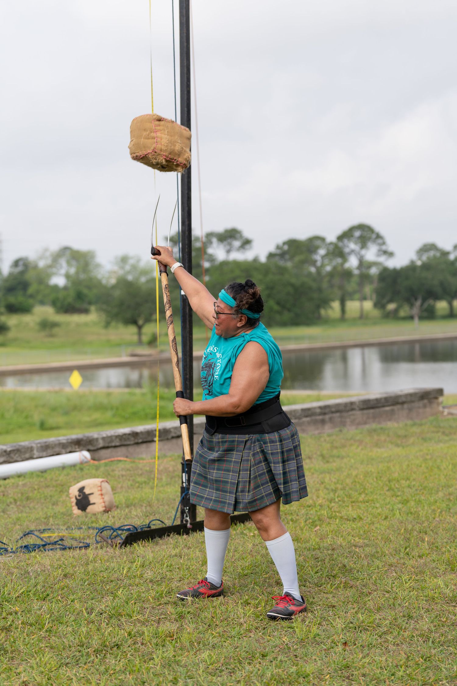 Woman in kilt and teal vest competes in Highland Games pole event on grassy field.