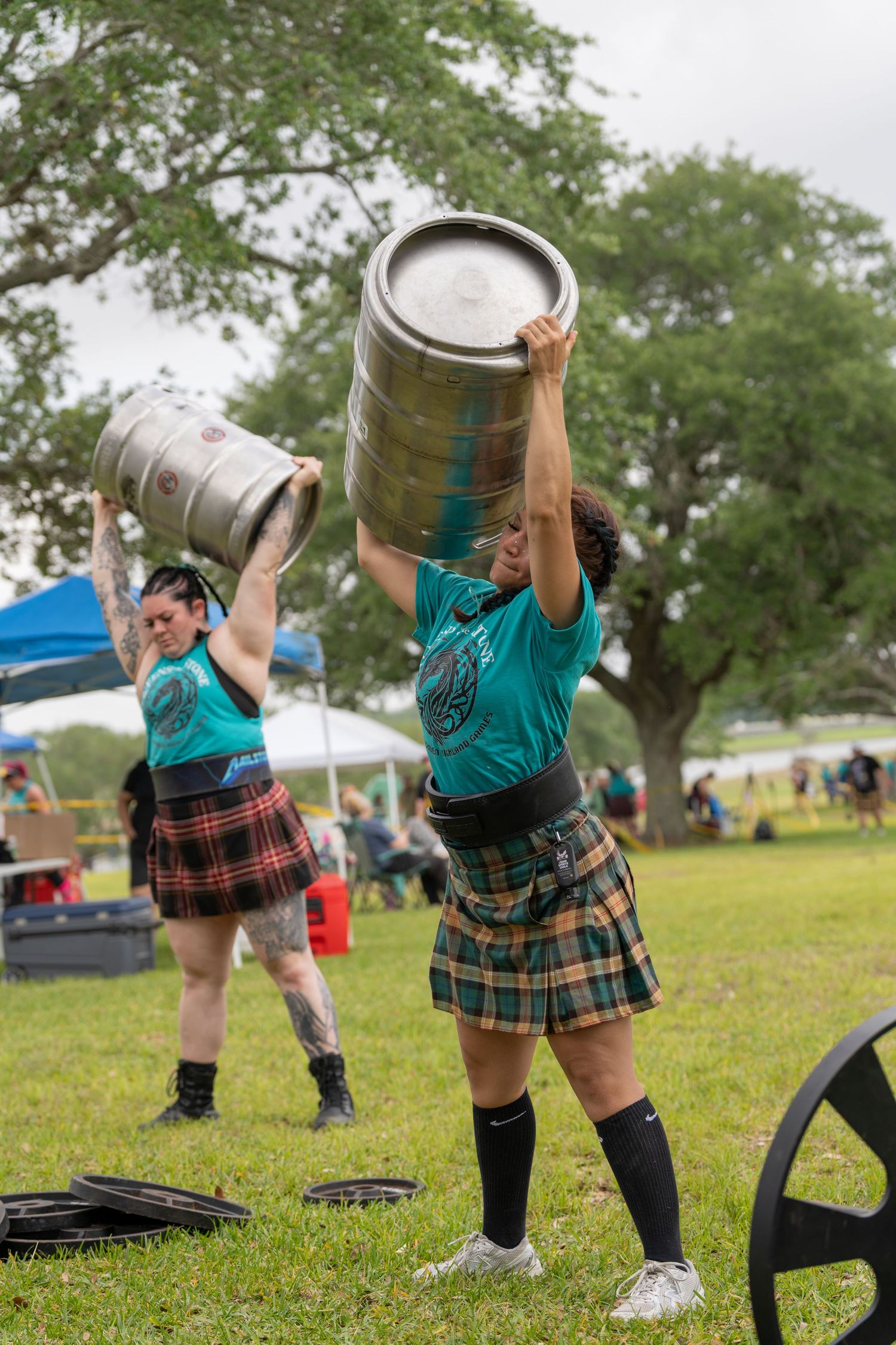 Two women in kilts lift metal kegs overhead at an outdoor Highland Games competition.