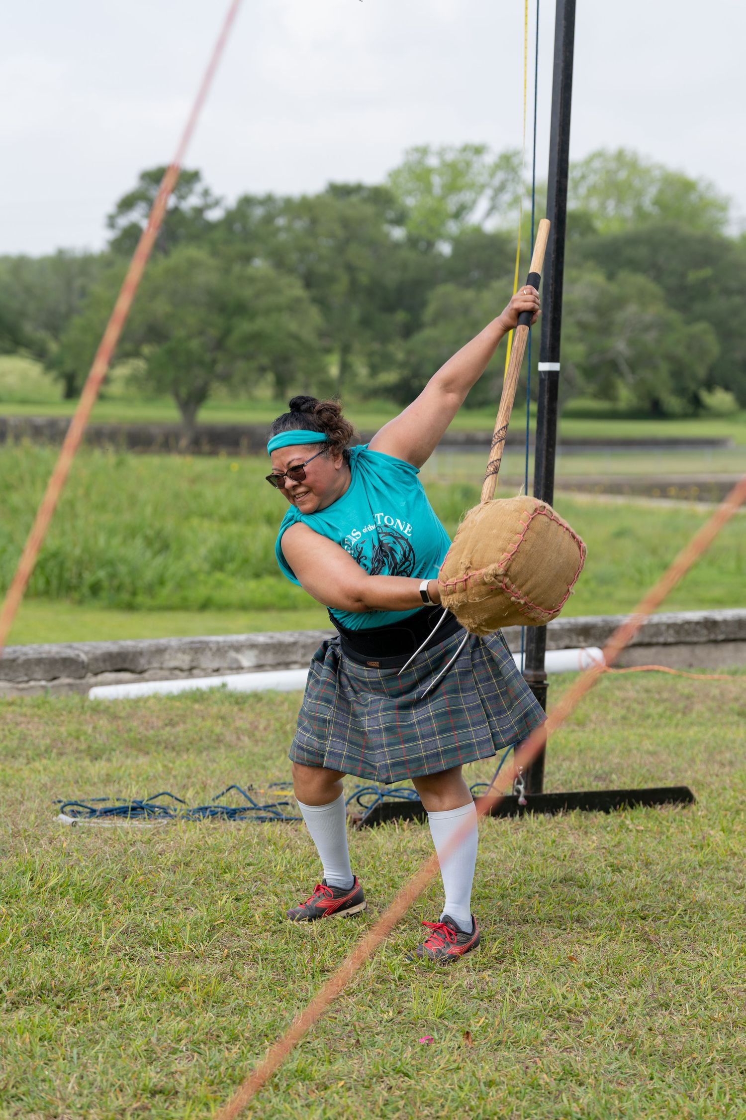 Woman in tartan kilt and teal shirt competes in Highland Games weight throw event on green grass field.