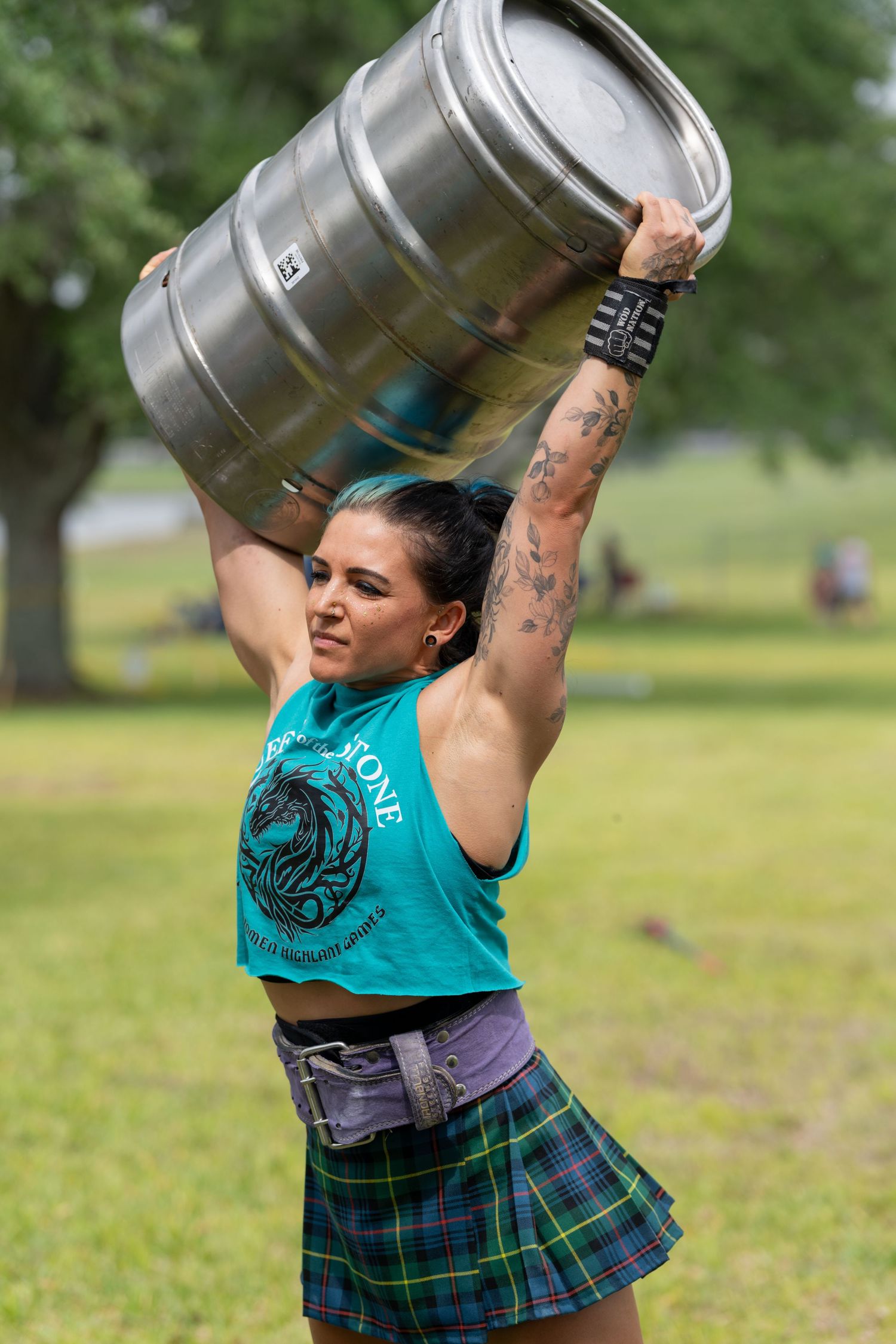 Tattooed woman in teal tank and plaid skirt lifts a metal keg overhead at an outdoor Highland games event.