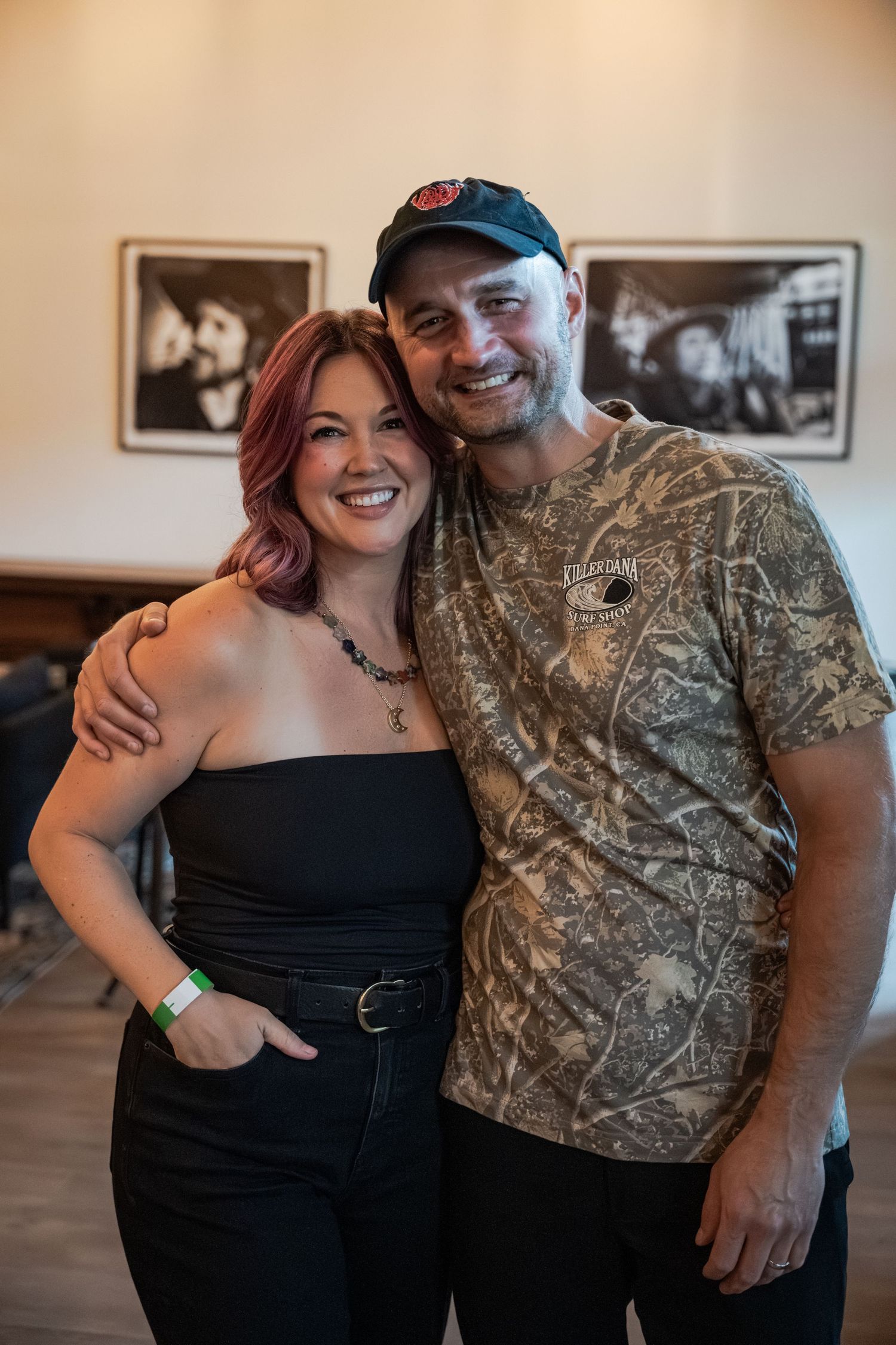 A smiling woman with red hair in a black top poses with a man in a camo shirt and black cap indoors.