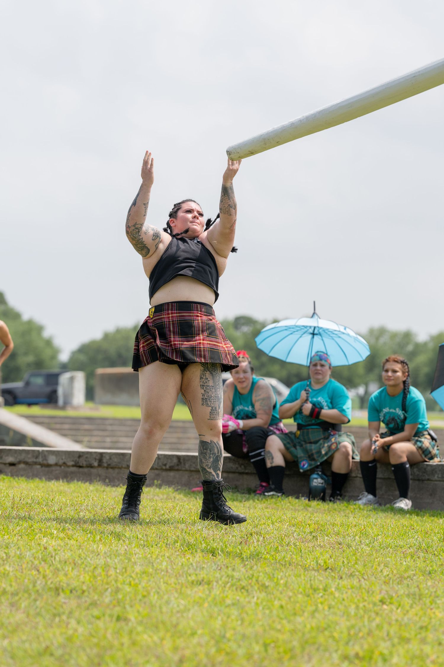 Woman in tartan kilt tosses caber at Highland Games competition while spectators watch from the sideline.