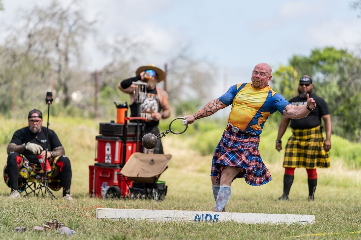 Man in tartan kilt competes in Highland Games throwing event outdoors, with spectators watching nearby.