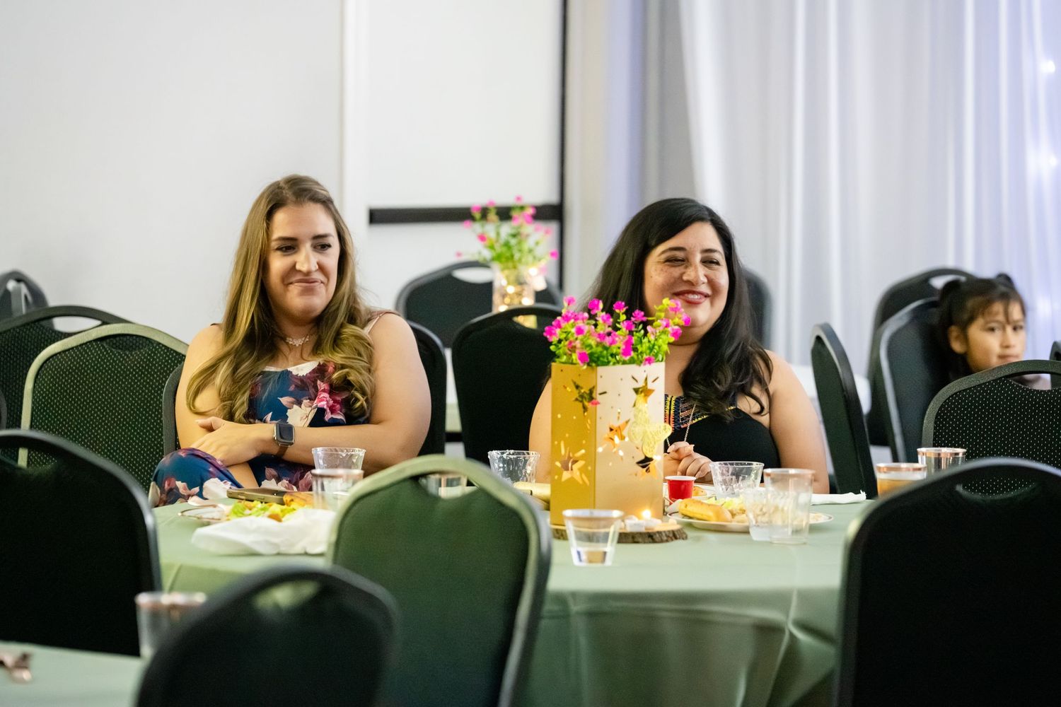 Two women smiling at a decorated banquet table with floral centerpieces at a formal event.