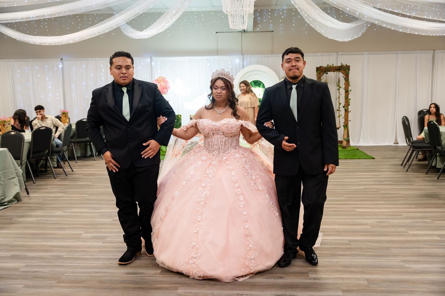 Young woman in pink quinceañera ball gown with crown, escorted by two men in black suits at formal celebration.