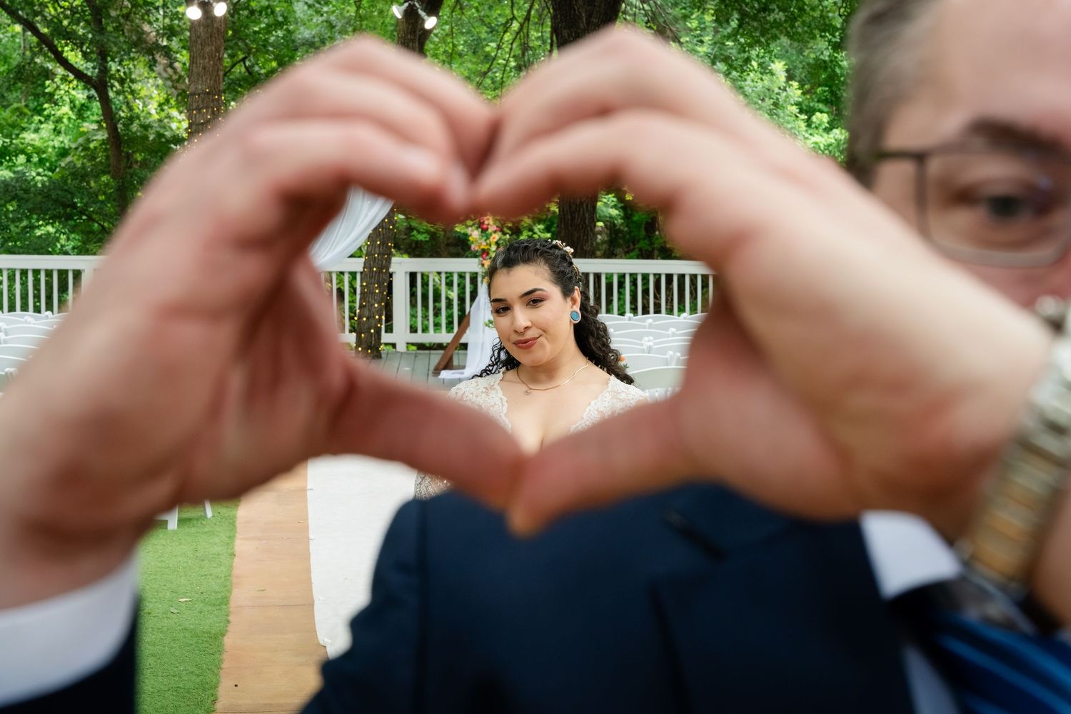 A person forms a heart shape with their hands framing a smiling bride in a white dress at an outdoor wedding venue.