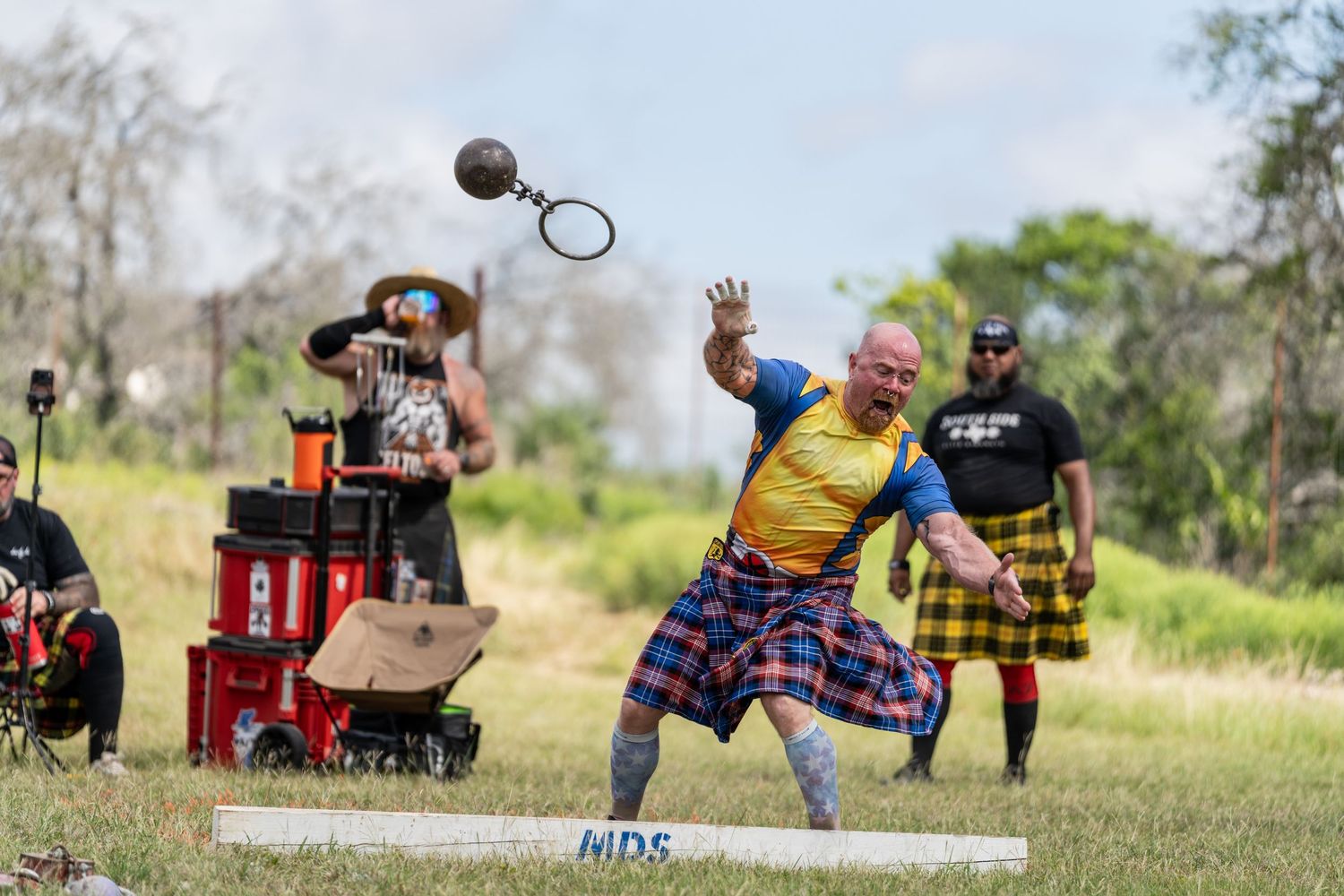 Man in tartan kilt throws weight over bar at Highland Games competition outdoors.
