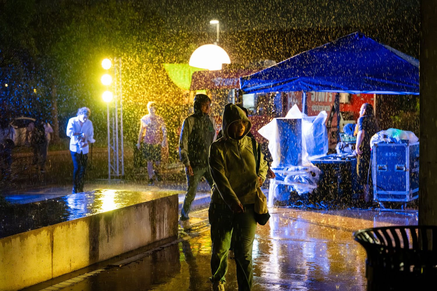 People walking in heavy rain at night near illuminated blue tent and street lights on wet pavement.