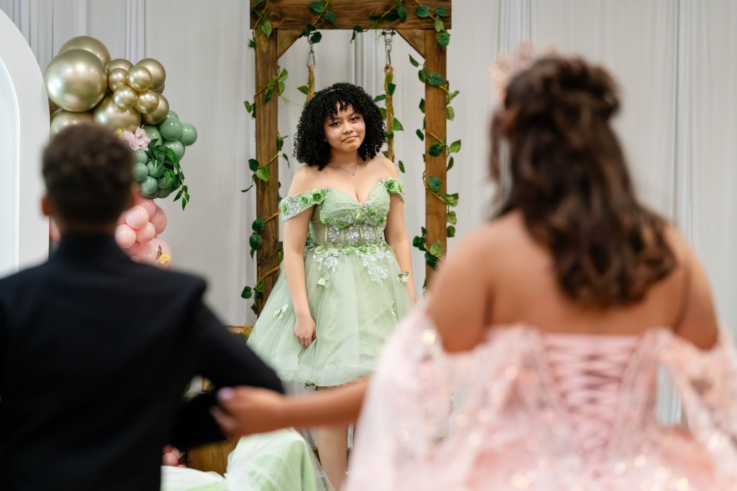 Young woman in green floral ball gown admires herself in mirror at quinceañera dress fitting.