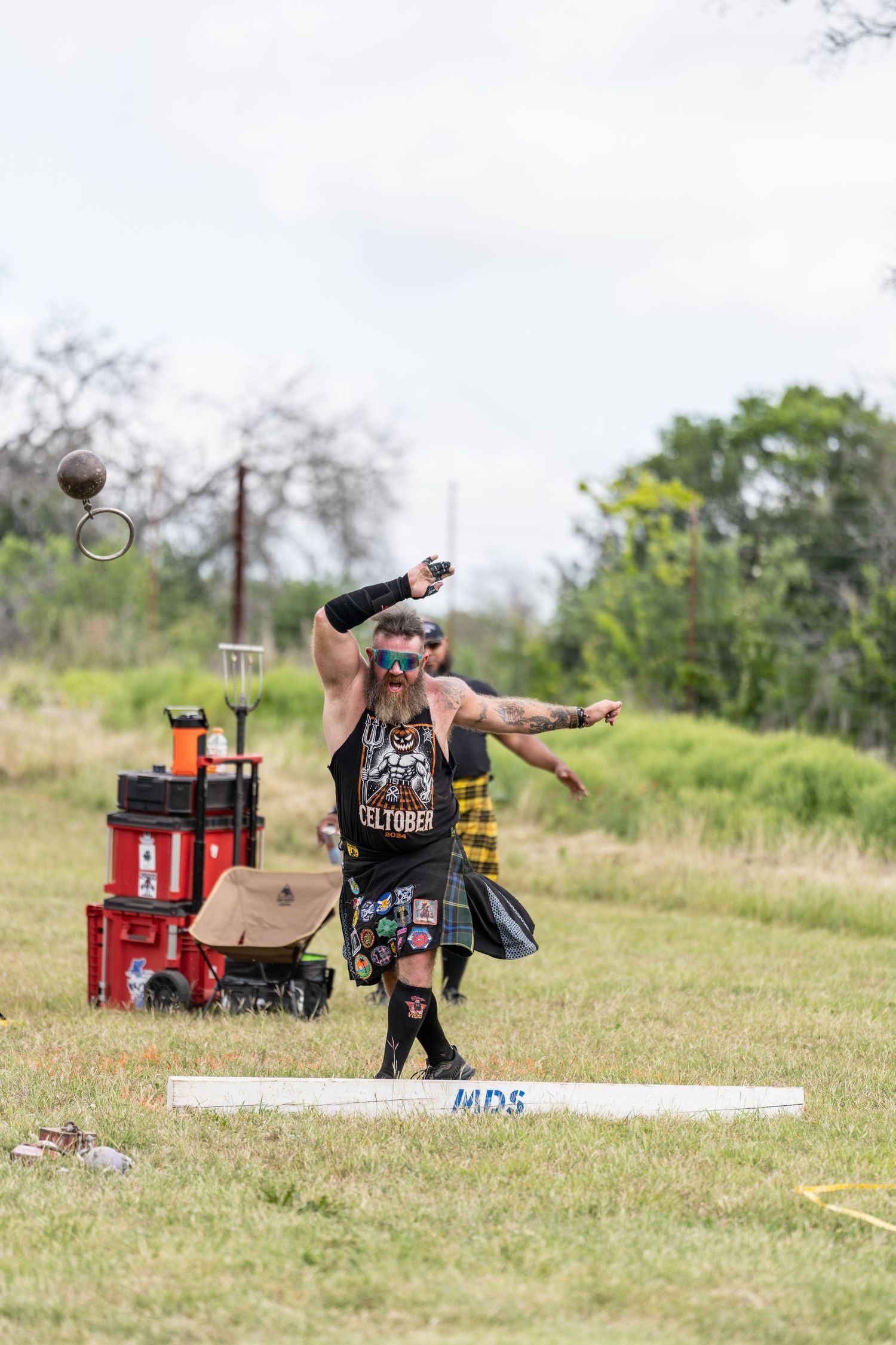 Man in kilt throwing shot put at Highland Games competition outdoors.