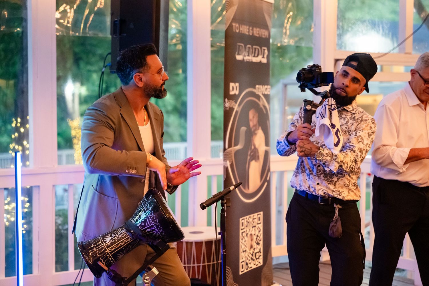 Two men at an event, one holding a camera on a gimbal stabilizer, the other gesturing while holding a decorative bag.
