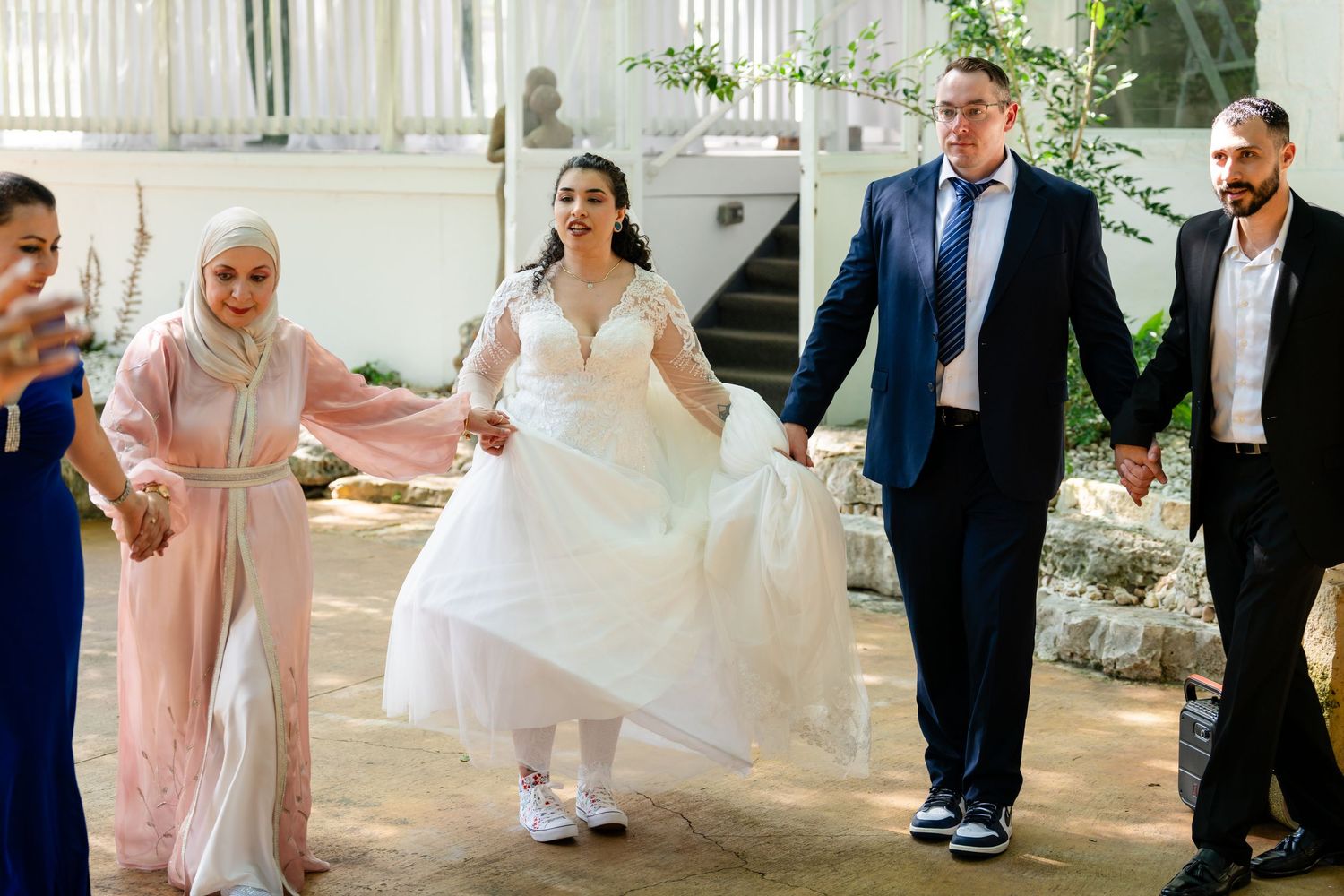 Bride in white gown holds hands with family members in elegant attire during a joyful outdoor wedding celebration.
