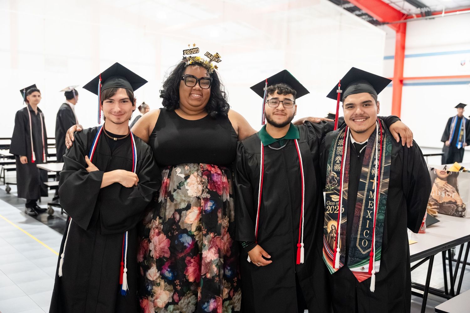 Four college graduates in caps and gowns smile together at an indoor graduation ceremony celebration.