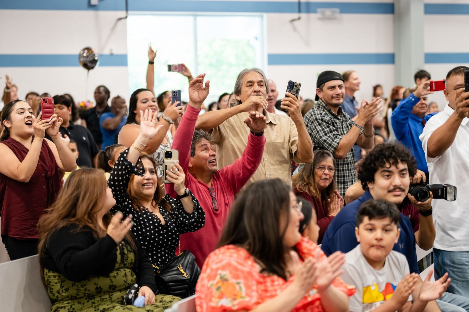 Diverse crowd of excited people clapping and taking photos with smartphones at an indoor event or celebration.