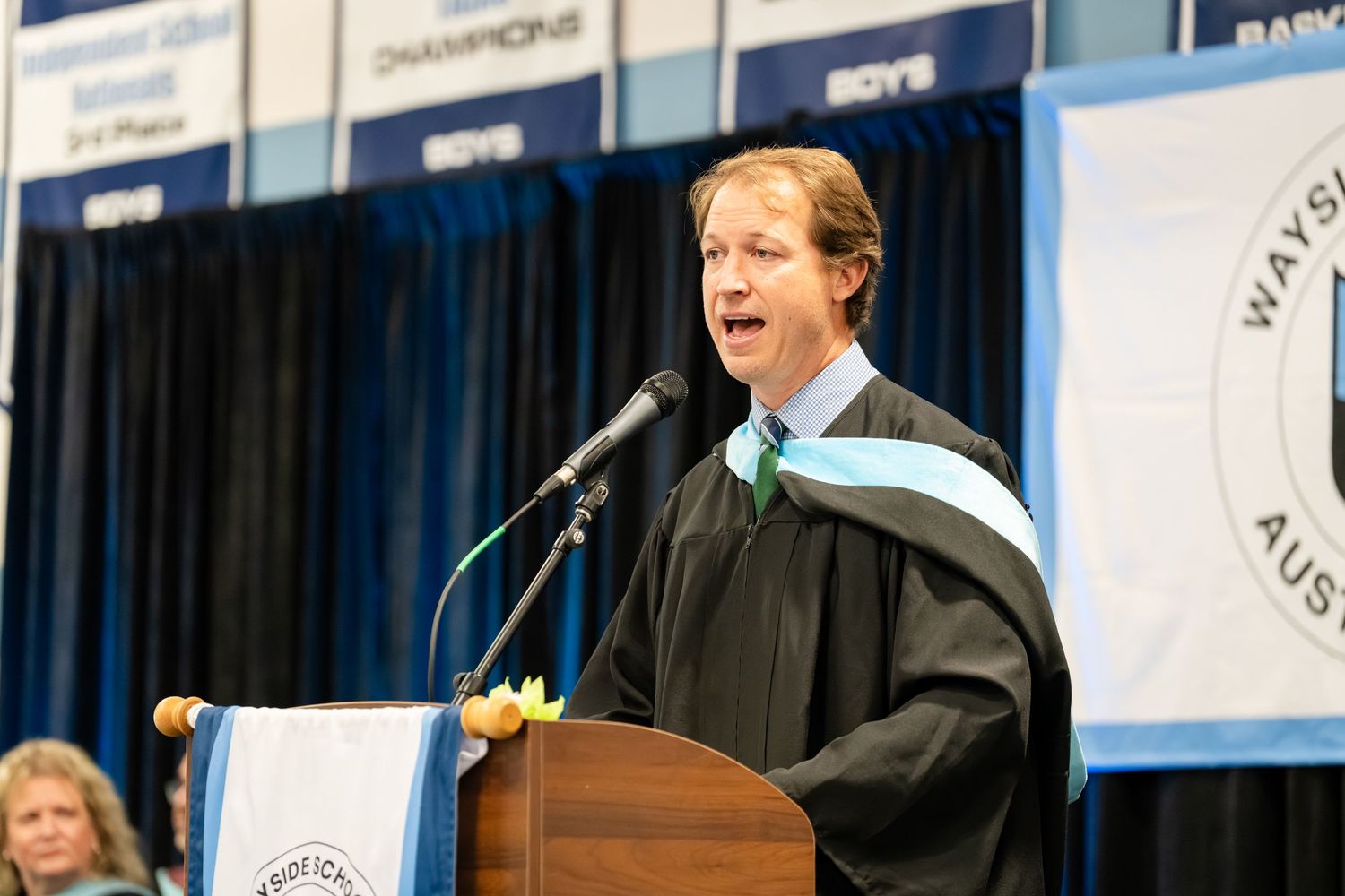 Man in academic regalia speaks at podium during graduation ceremony in gymnasium with championship banners.