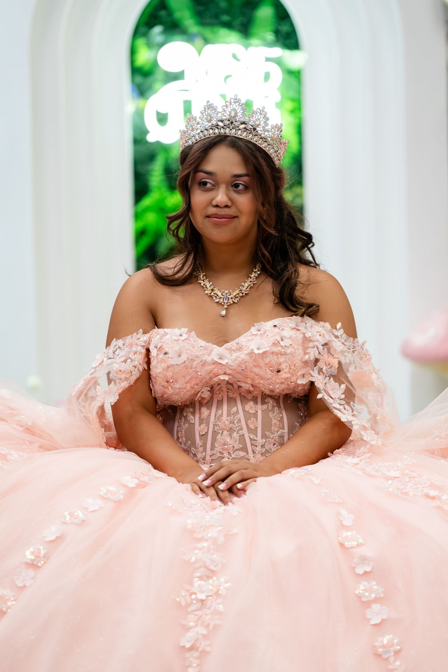 Young woman in pink quinceañera ball gown with floral lace, diamond crown and necklace, posing elegantly indoors.