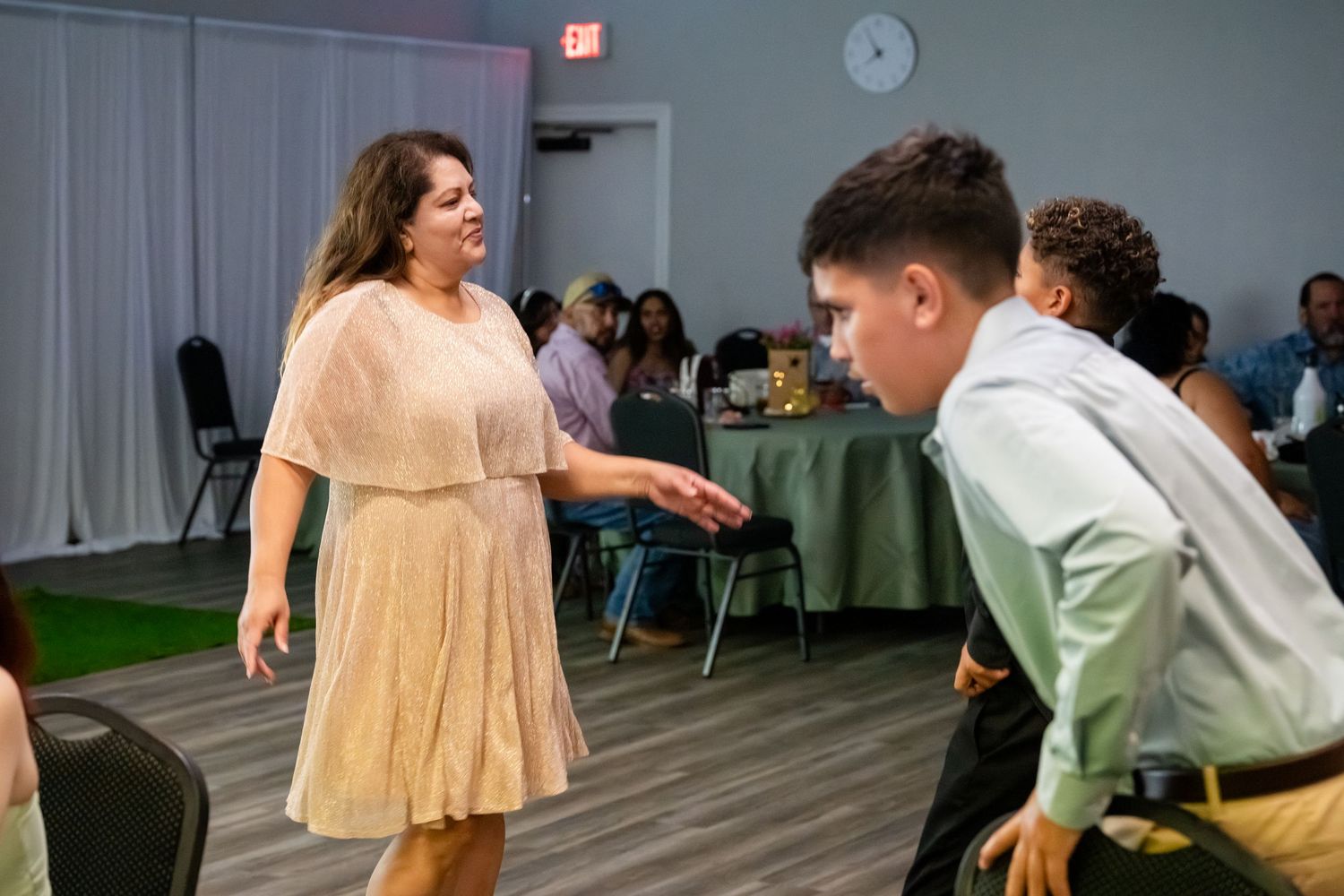 Woman in beige dress dancing with young man in green shirt at an indoor celebration event.