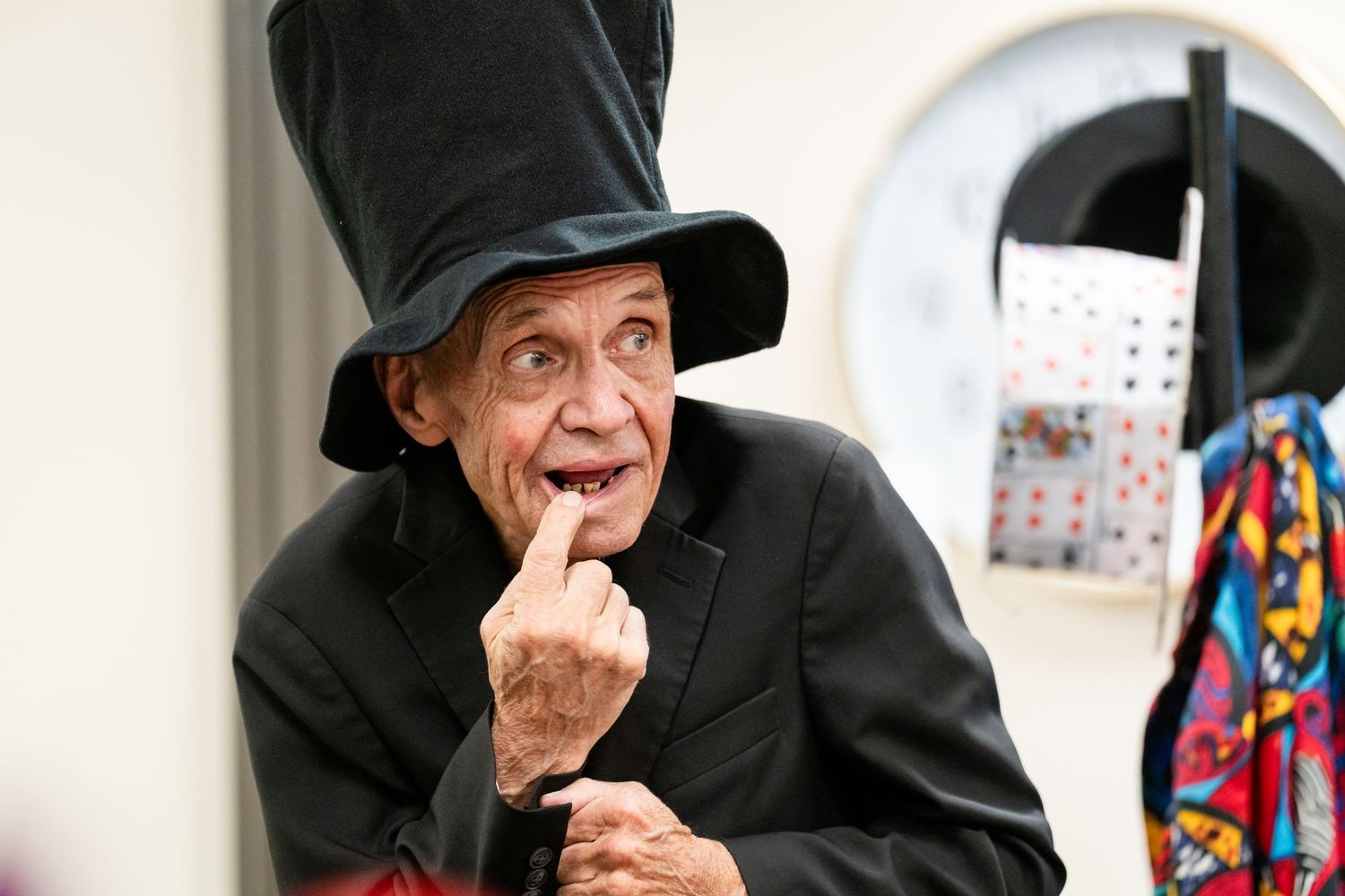 Elderly man in tall black top hat and dark suit gesturing with finger, performing as a magician or entertainer.