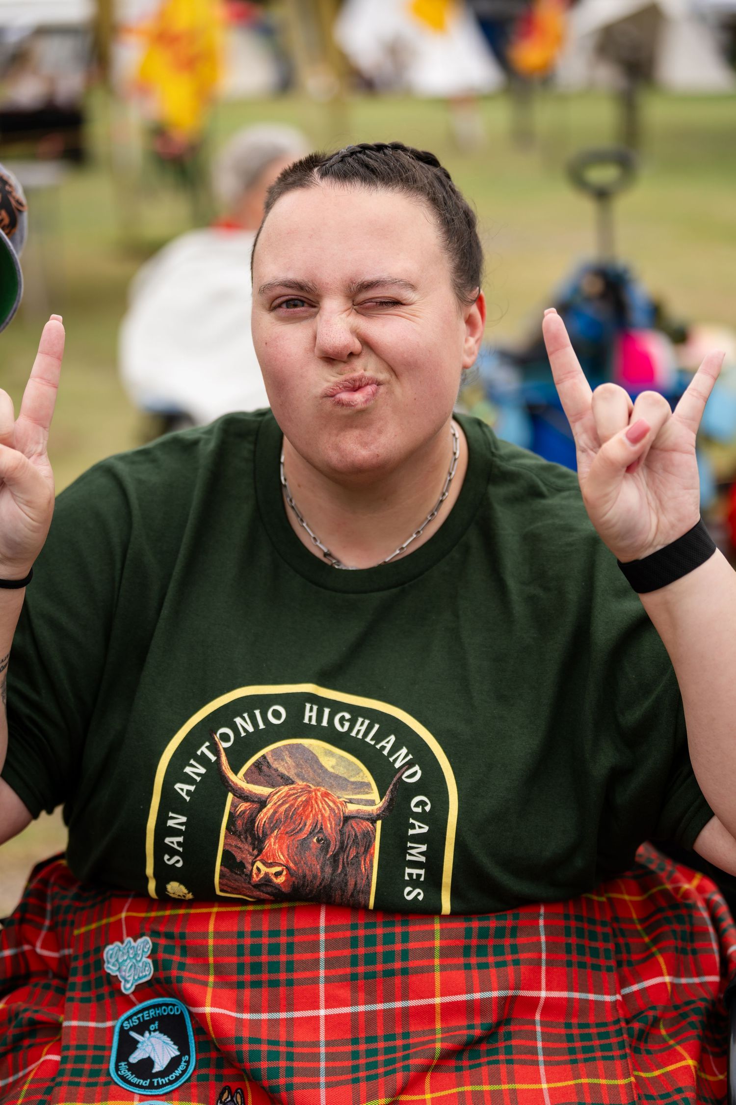 Person wearing San Antonio Highland Games shirt making rock on hand gestures at an outdoor Scottish festival event.