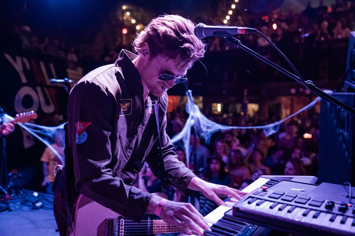 Musician performs on keyboard at concert venue with dramatic blue stage lighting and crowd in background.