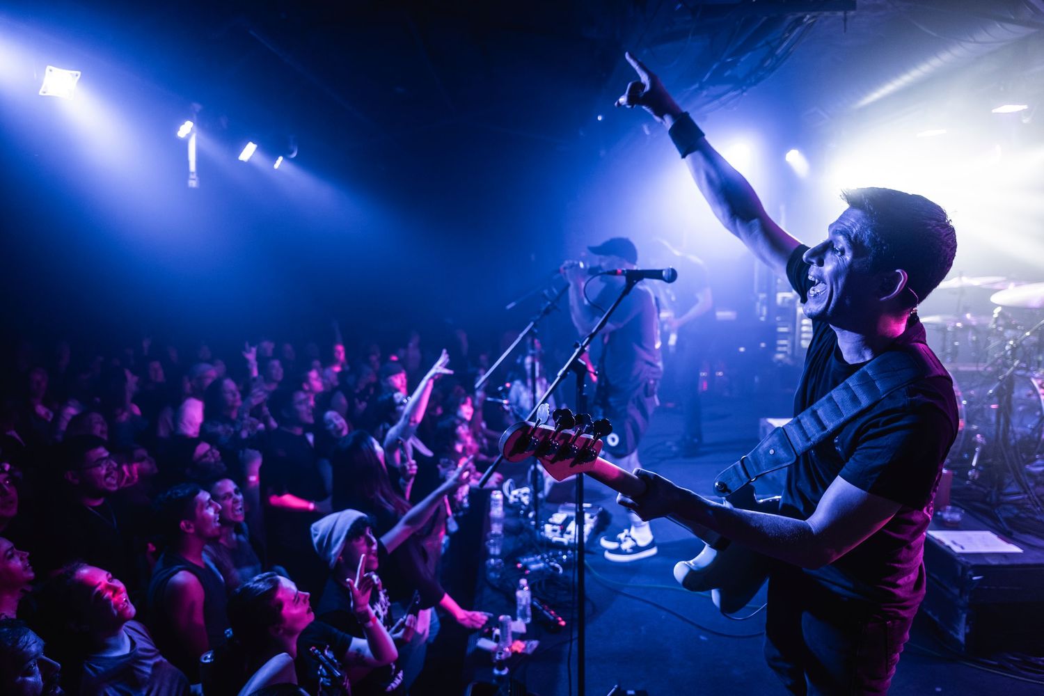 Musician performs on stage with guitar at concert venue with blue lighting and crowd raising hands in foreground.