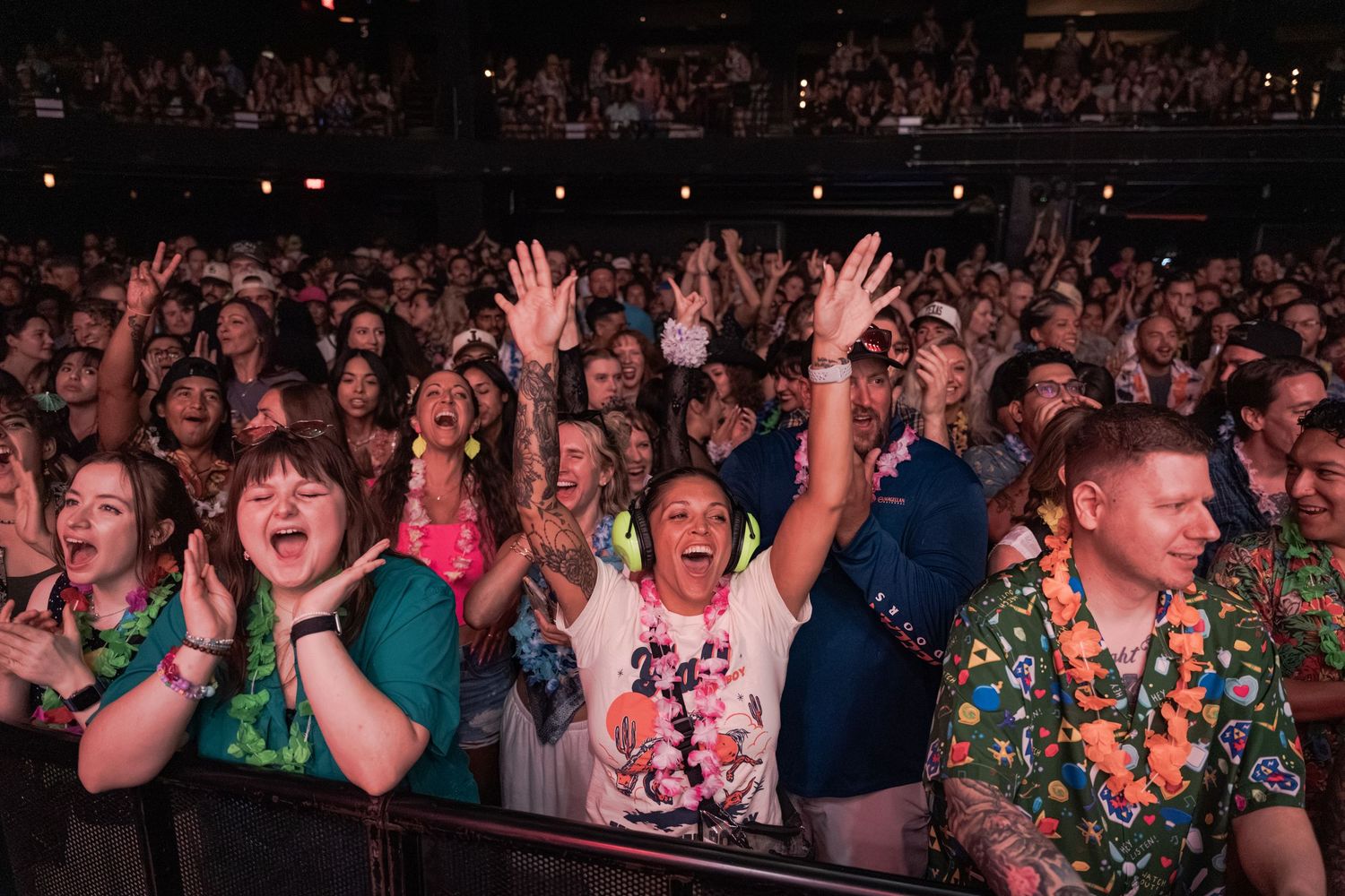 Excited concert crowd cheering with hands raised at a lively indoor music event, fans wearing colorful outfits.