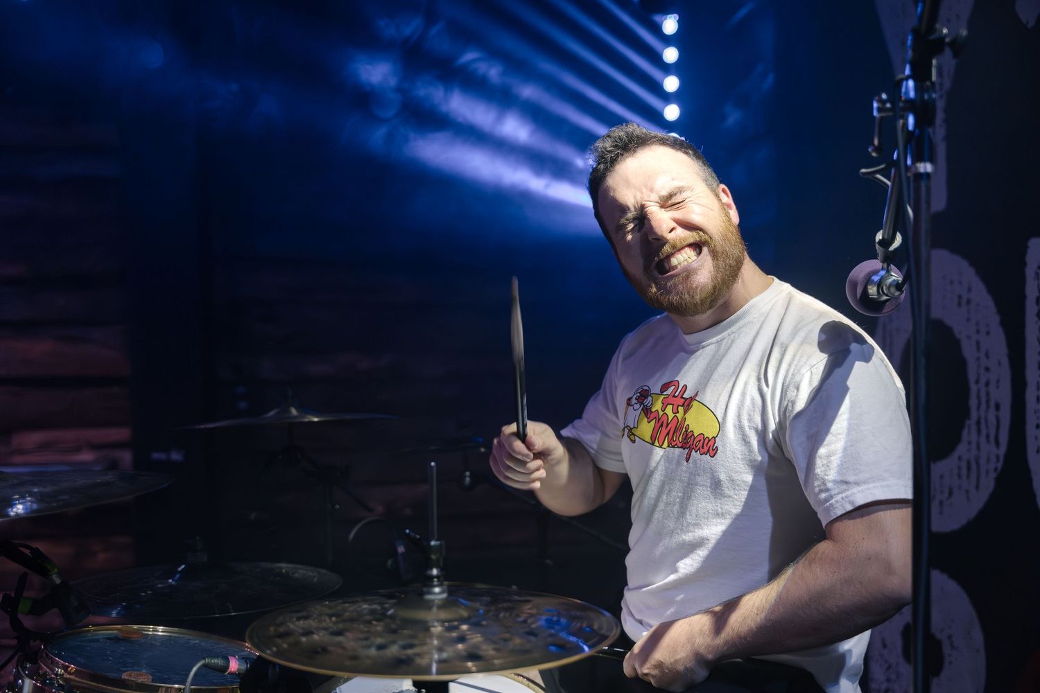 A drummer smiles while performing on stage with dramatic blue lighting effects.