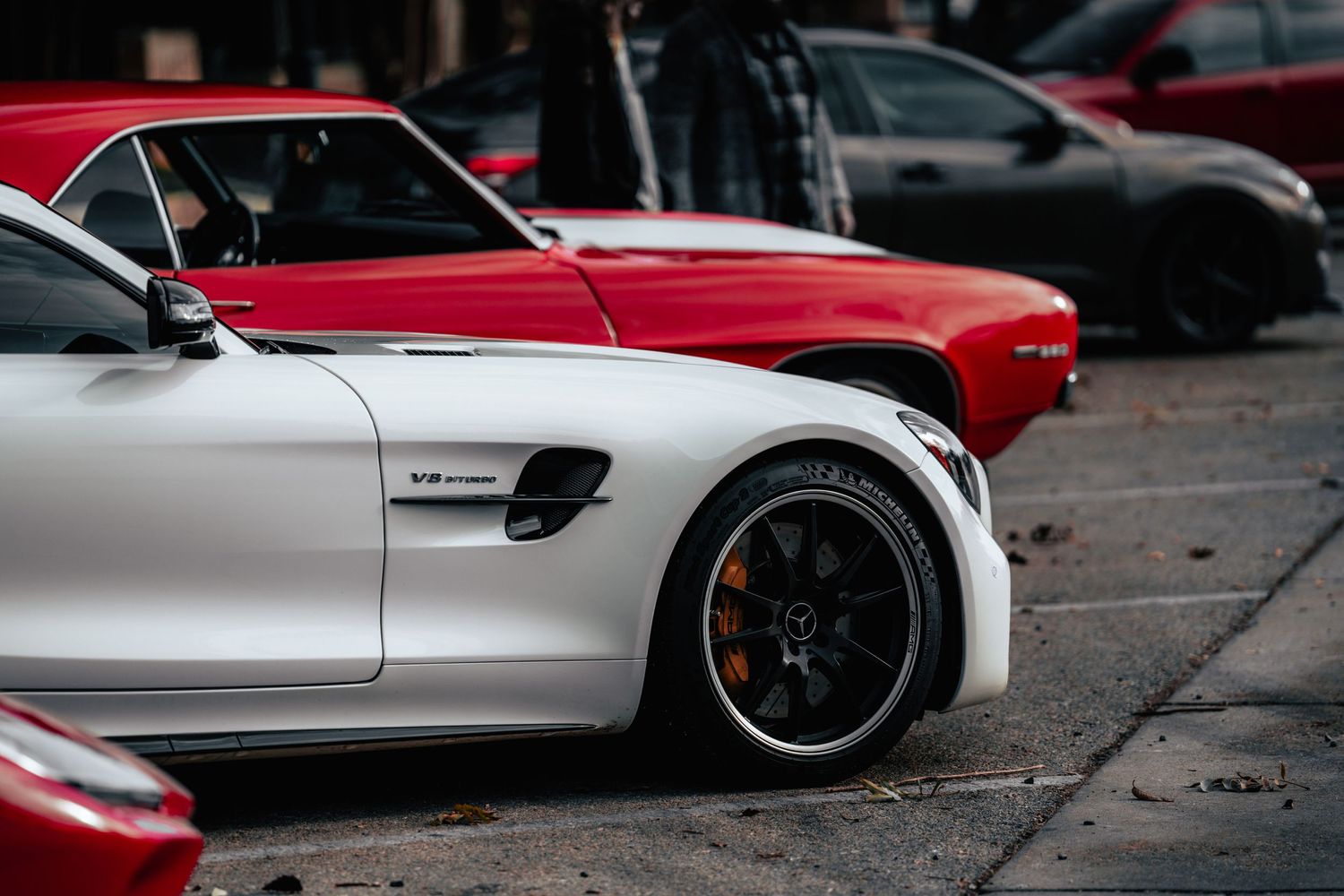 Close-up view of a silver Mercedes AMG sports car wheel and fender with a red car in the background.