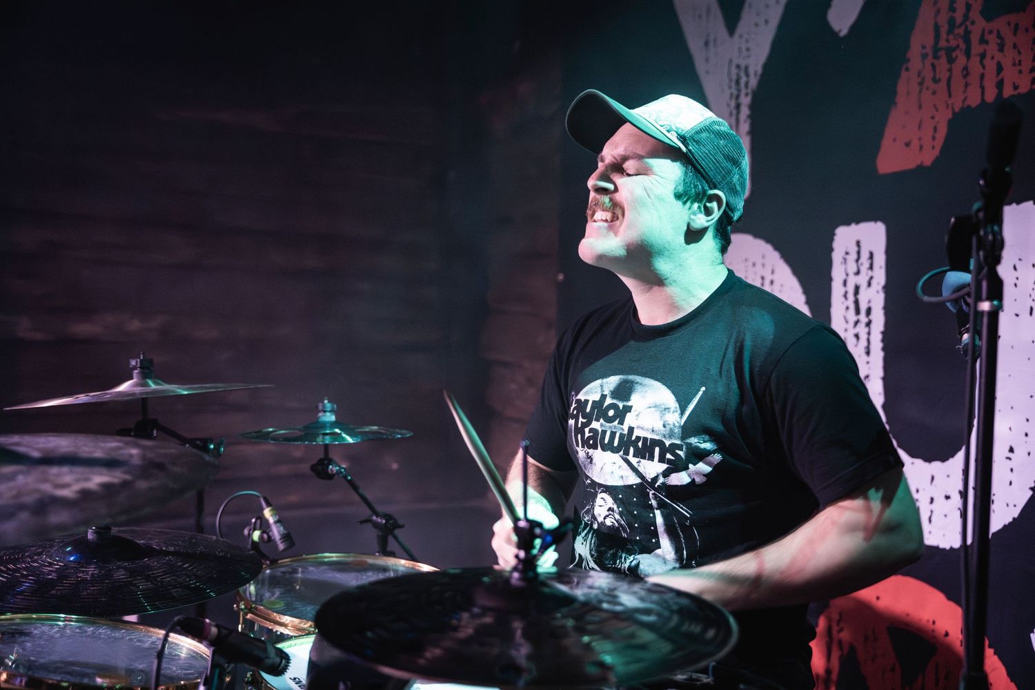 Energetic drummer wearing baseball cap performs during live concert under colored stage lights.