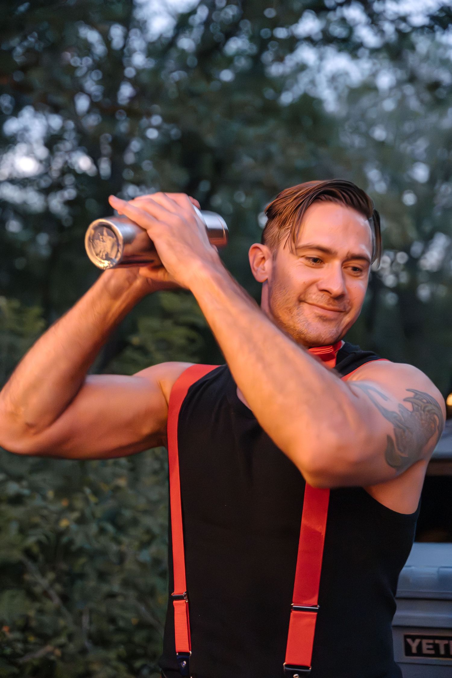 Person in black with red suspenders pouring a drink outdoors at twilight.