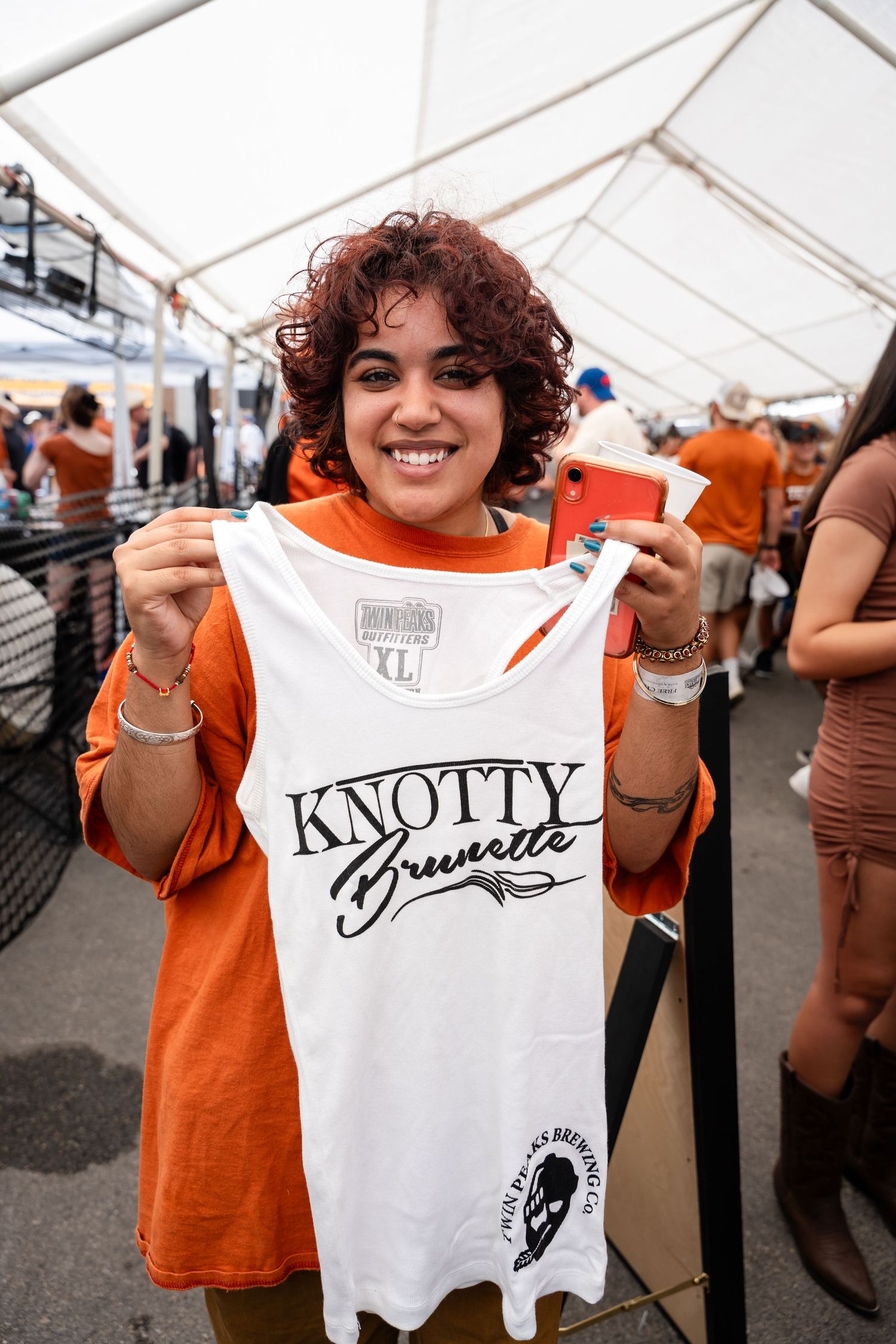Fan in orange shirt holding up Knotty Brunette branded merchandise at outdoor event.