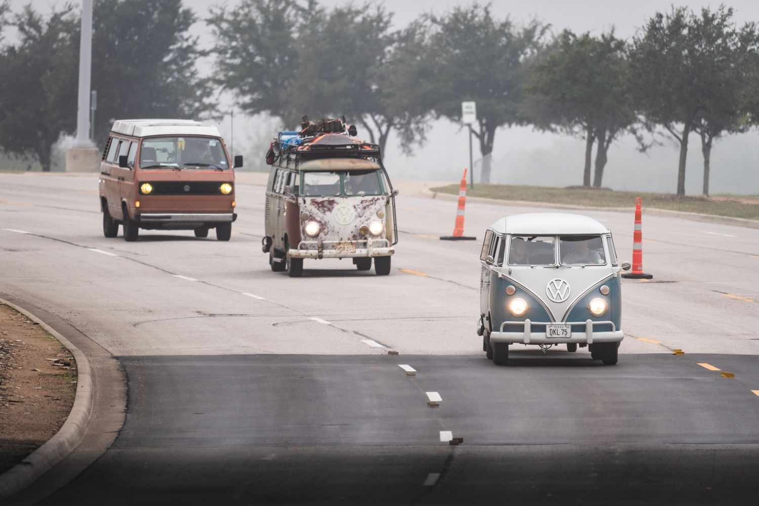 Classic VW bus and vintage vehicles drive along a misty road lined with trees and traffic cones.