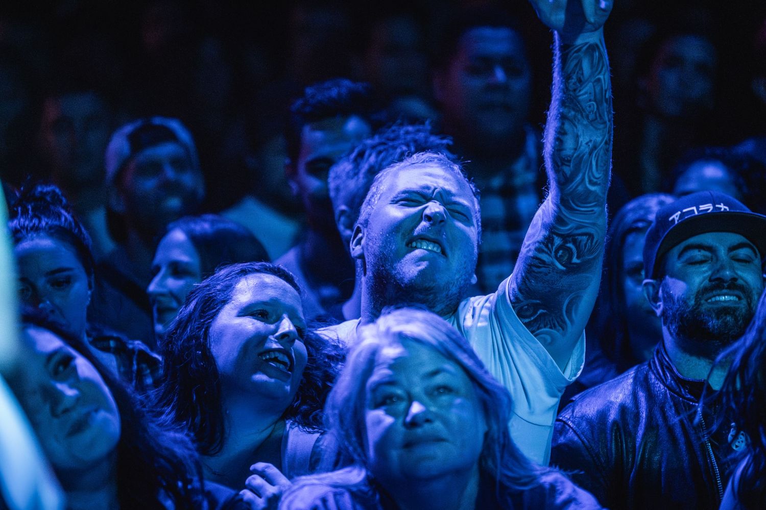 Concert crowd illuminated by blue stage lights with raised arms and enthusiastic expressions.