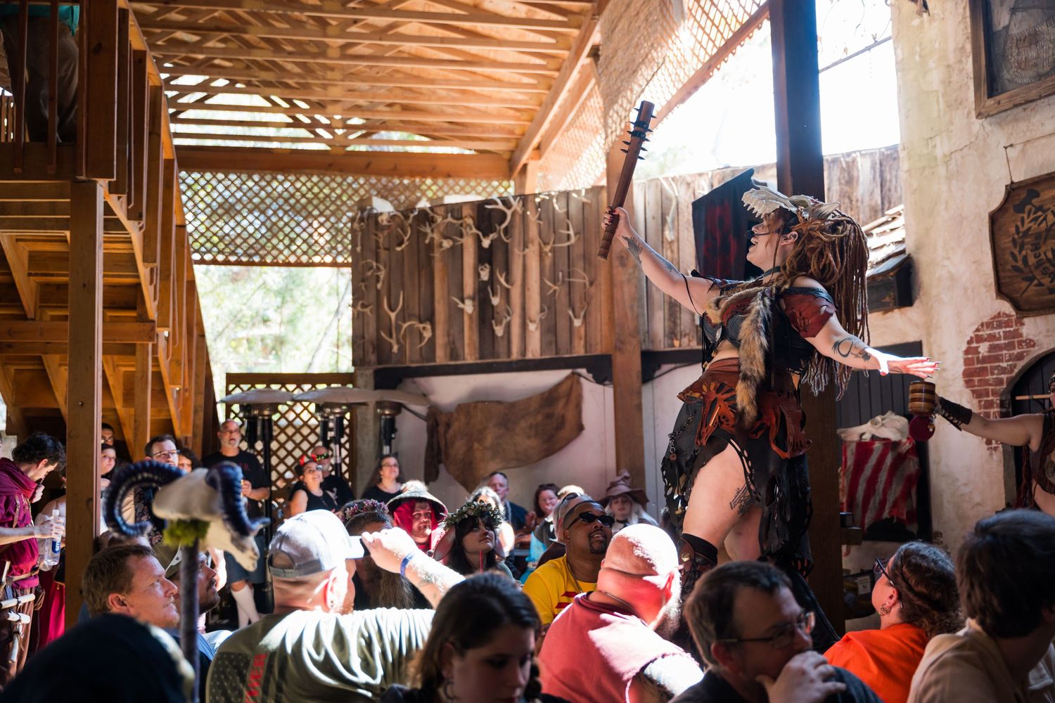 A crowded audience watches performers at a Renaissance festival under a wooden covered area.
