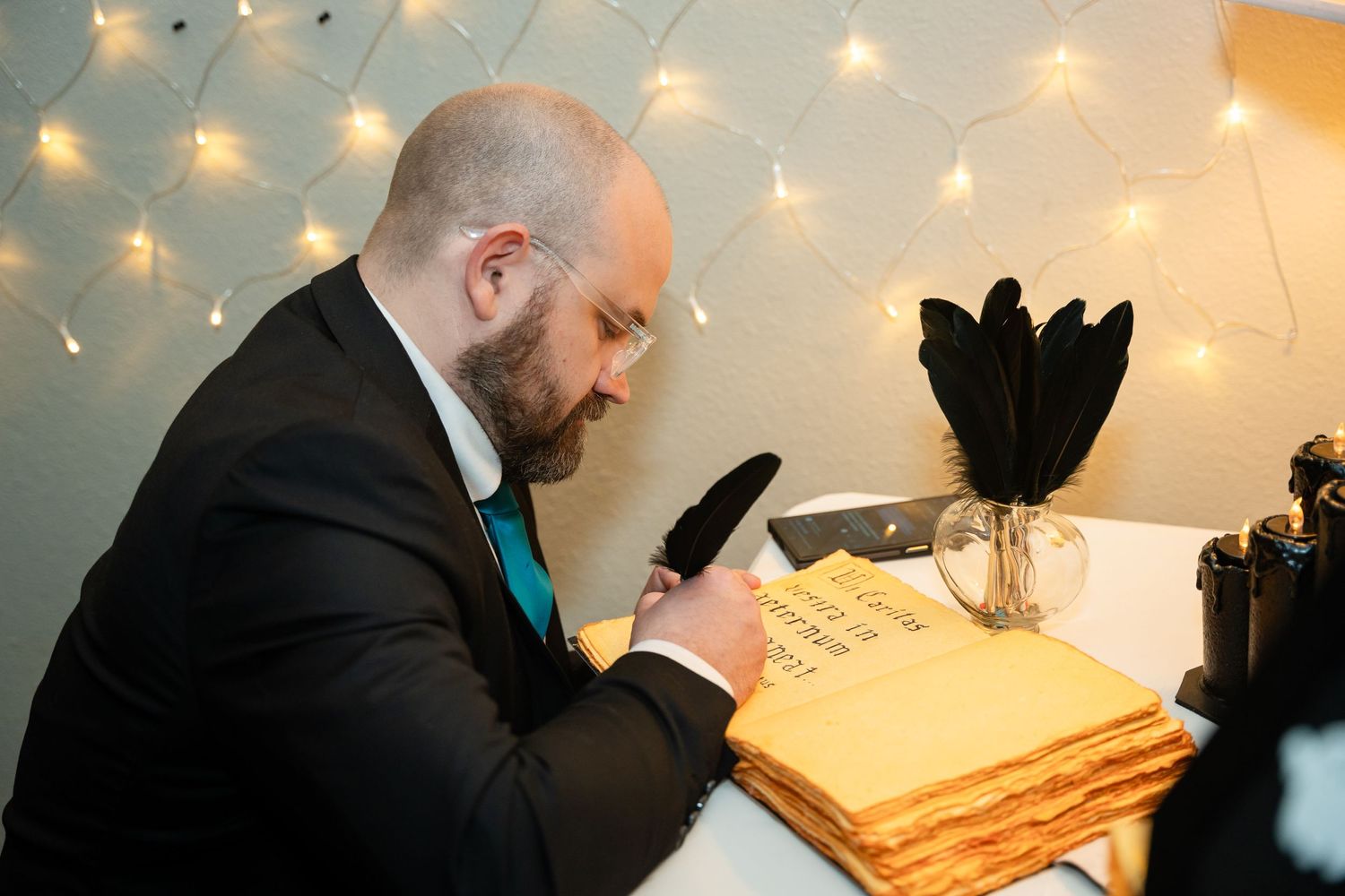 Person in formal attire signs guest book at decorated table with string lights overhead.