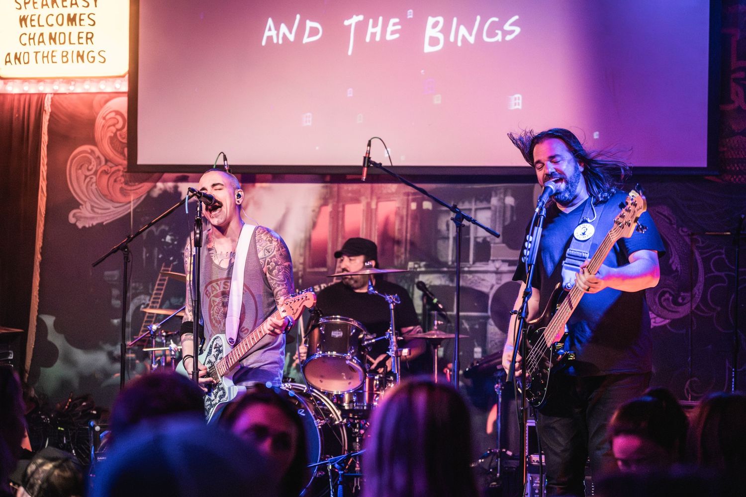 Three-piece band performs on stage with dramatic lighting and projection screen backdrop.
