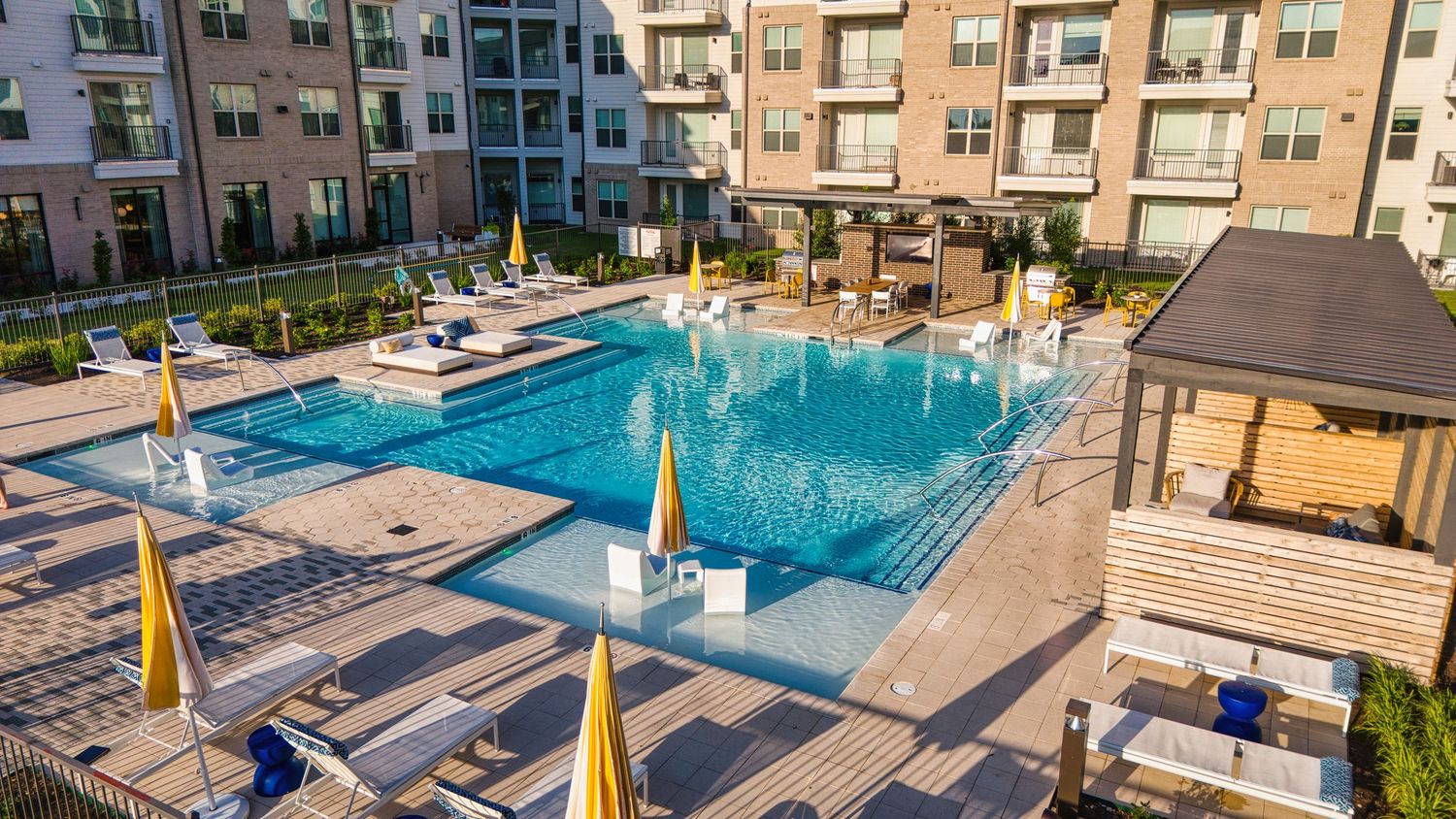Modern apartment complex with resort-style pool featuring yellow umbrellas and wooden pergolas.