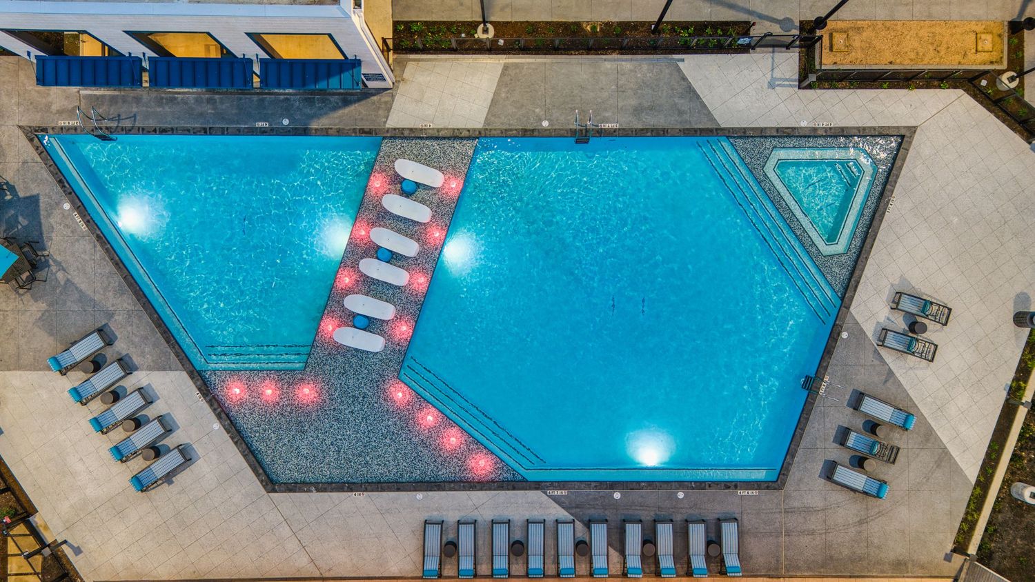Illuminated geometric swimming pool with loungers viewed from above at dusk.