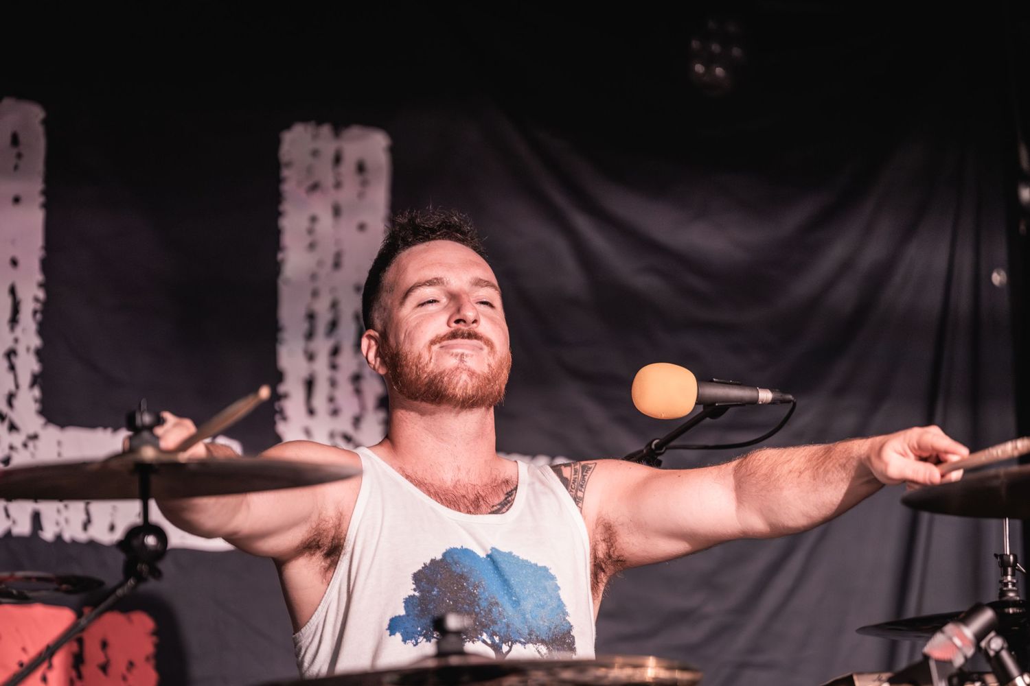 Drummer performs passionately on stage with arms spread wide under bright spotlights.