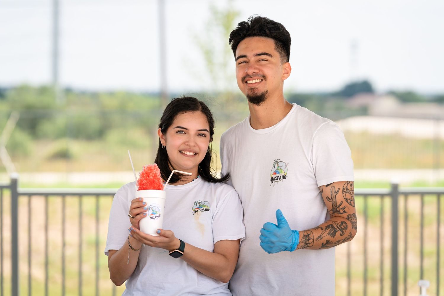 Two retail workers in white shirts hold beverages while posing outdoors.