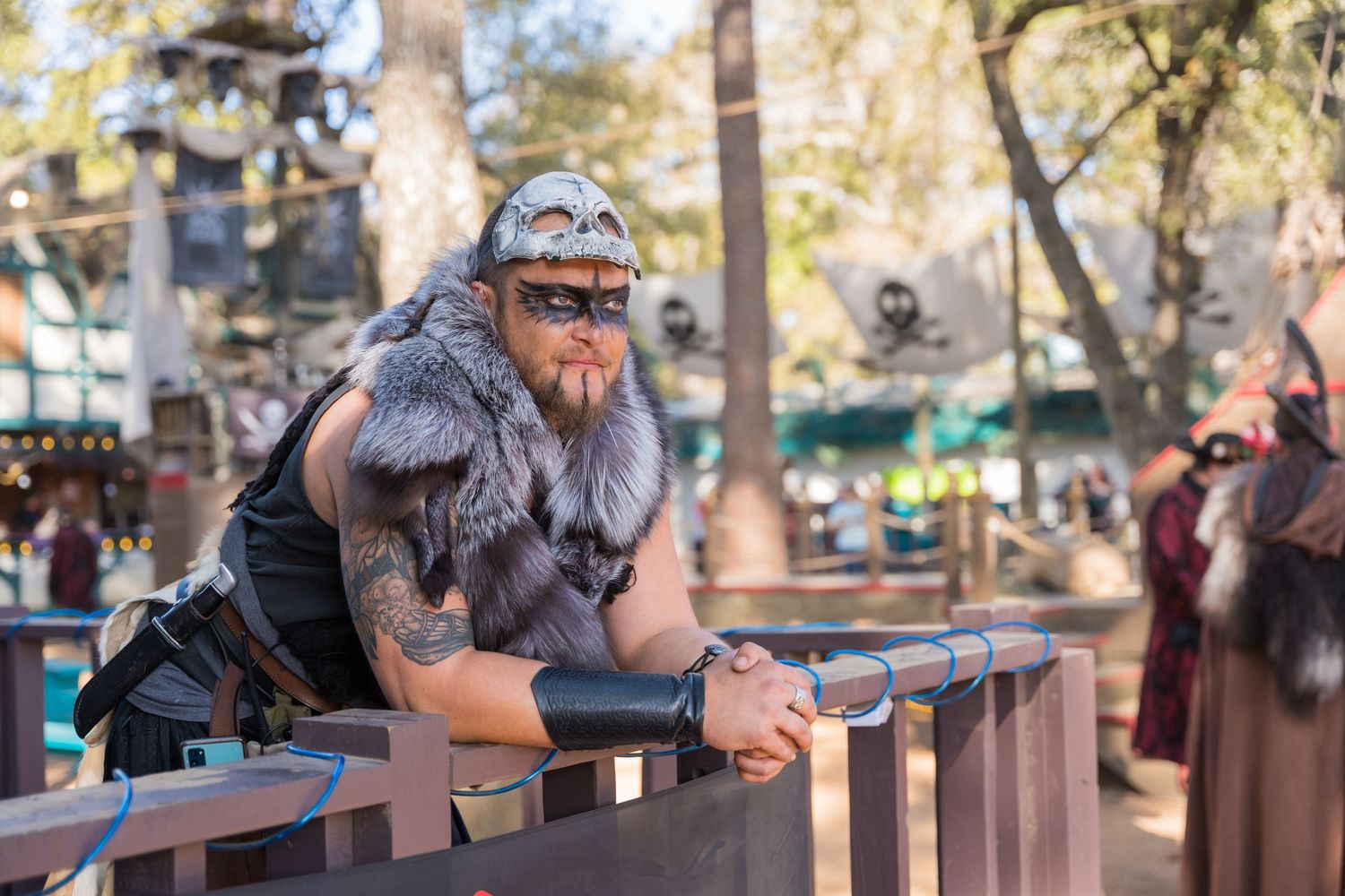 Costumed performer in fur and leather leans on wooden railing at renaissance festival.