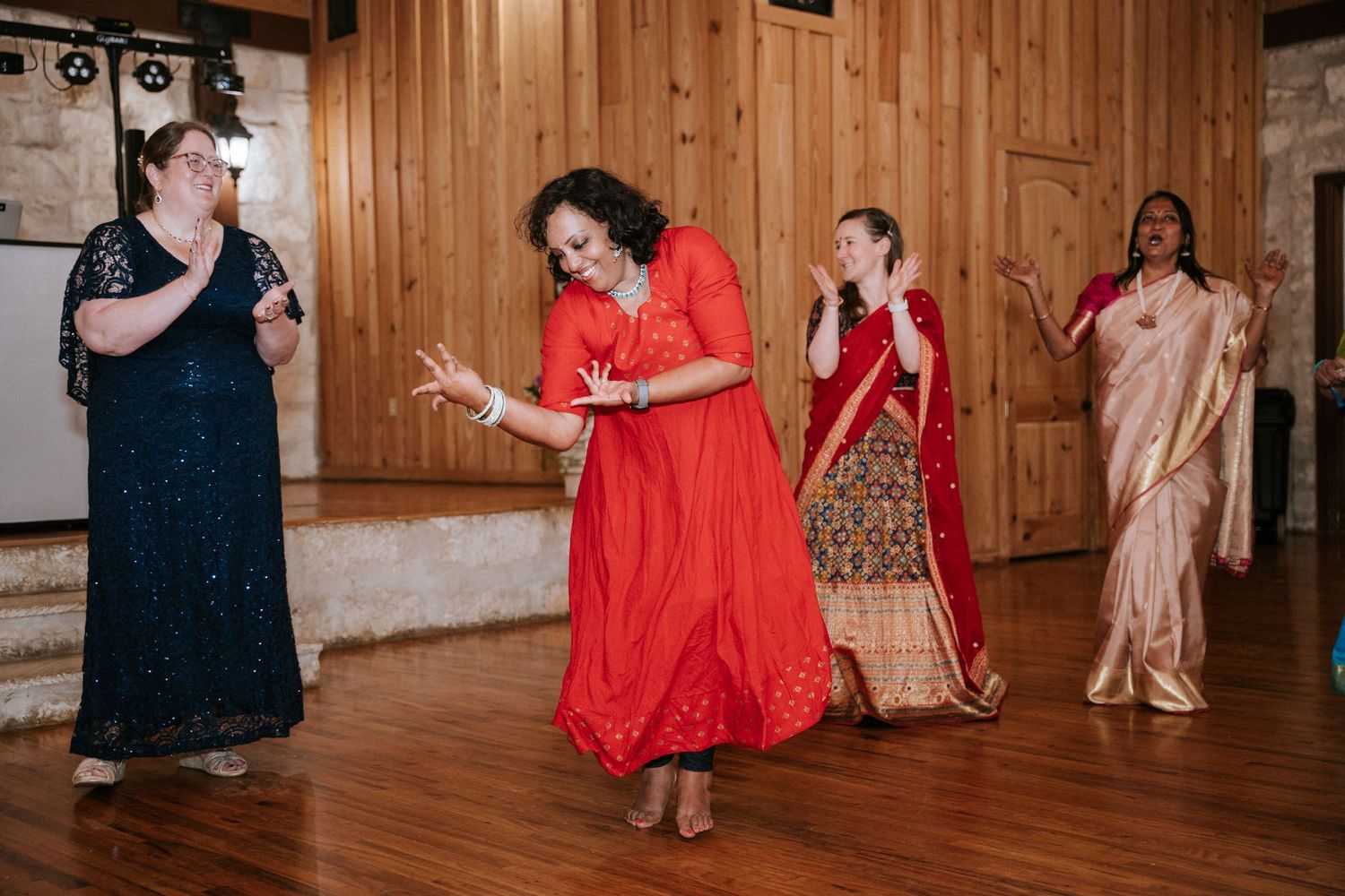Women in colorful dresses dance together at a celebration on a wooden dance floor.