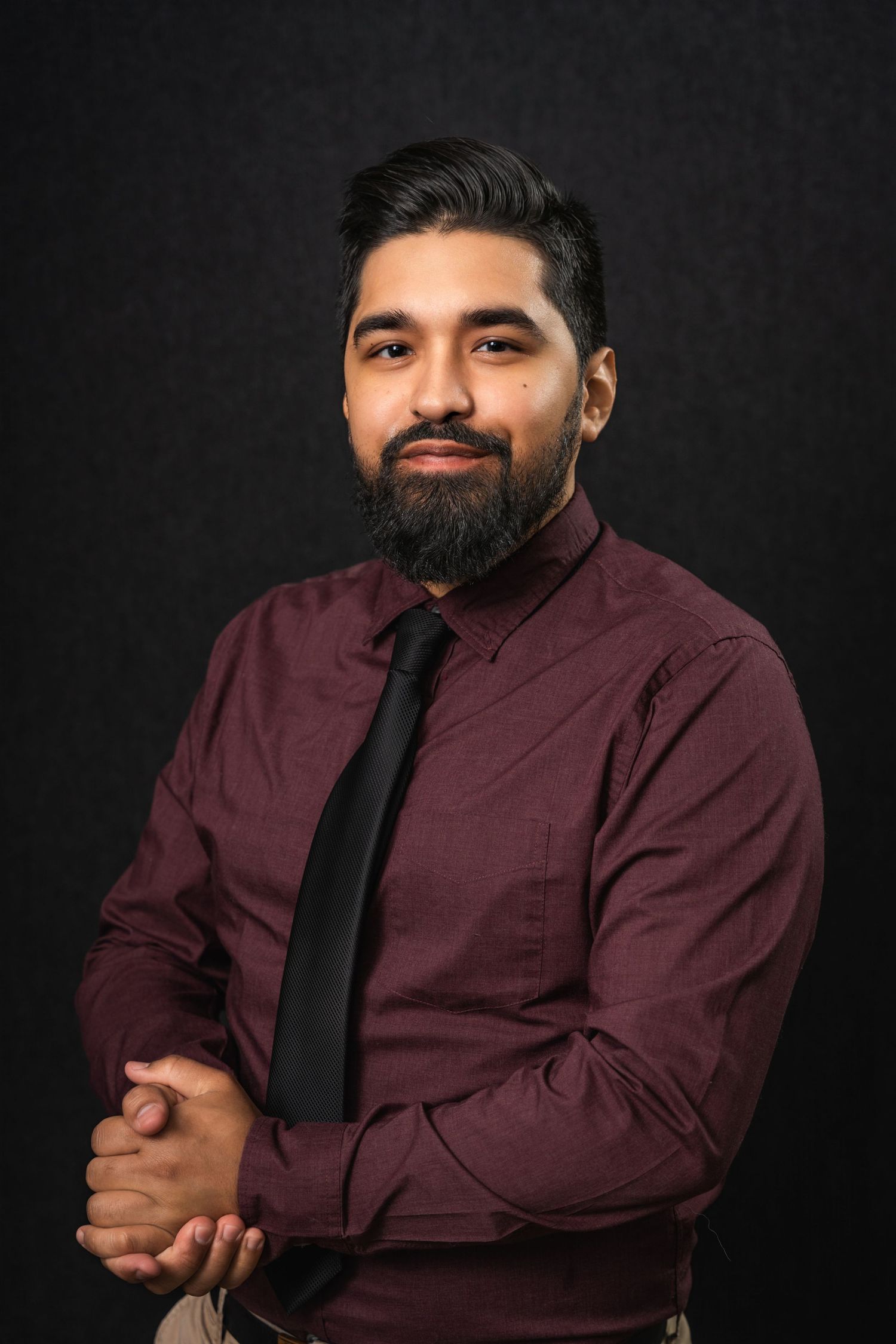 Professional headshot of person in burgundy dress shirt and black tie against dark backdrop.
