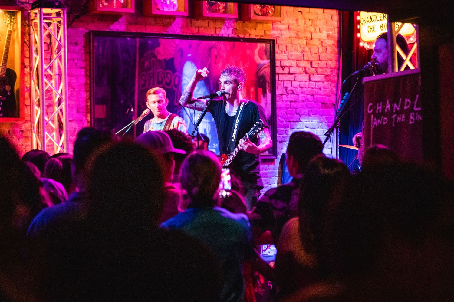 Band performing for crowd in intimate venue with purple lighting and brick walls.