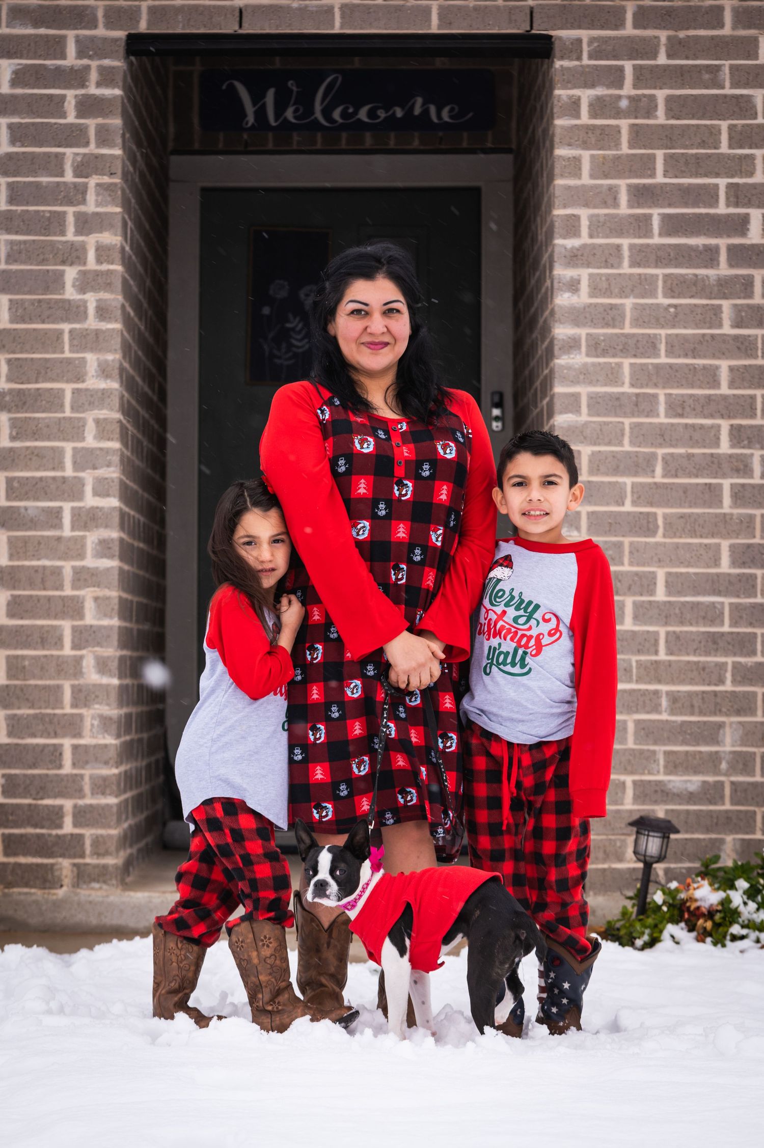 Family wearing matching red holiday outfits poses with their dog in front of a brick house in snow.