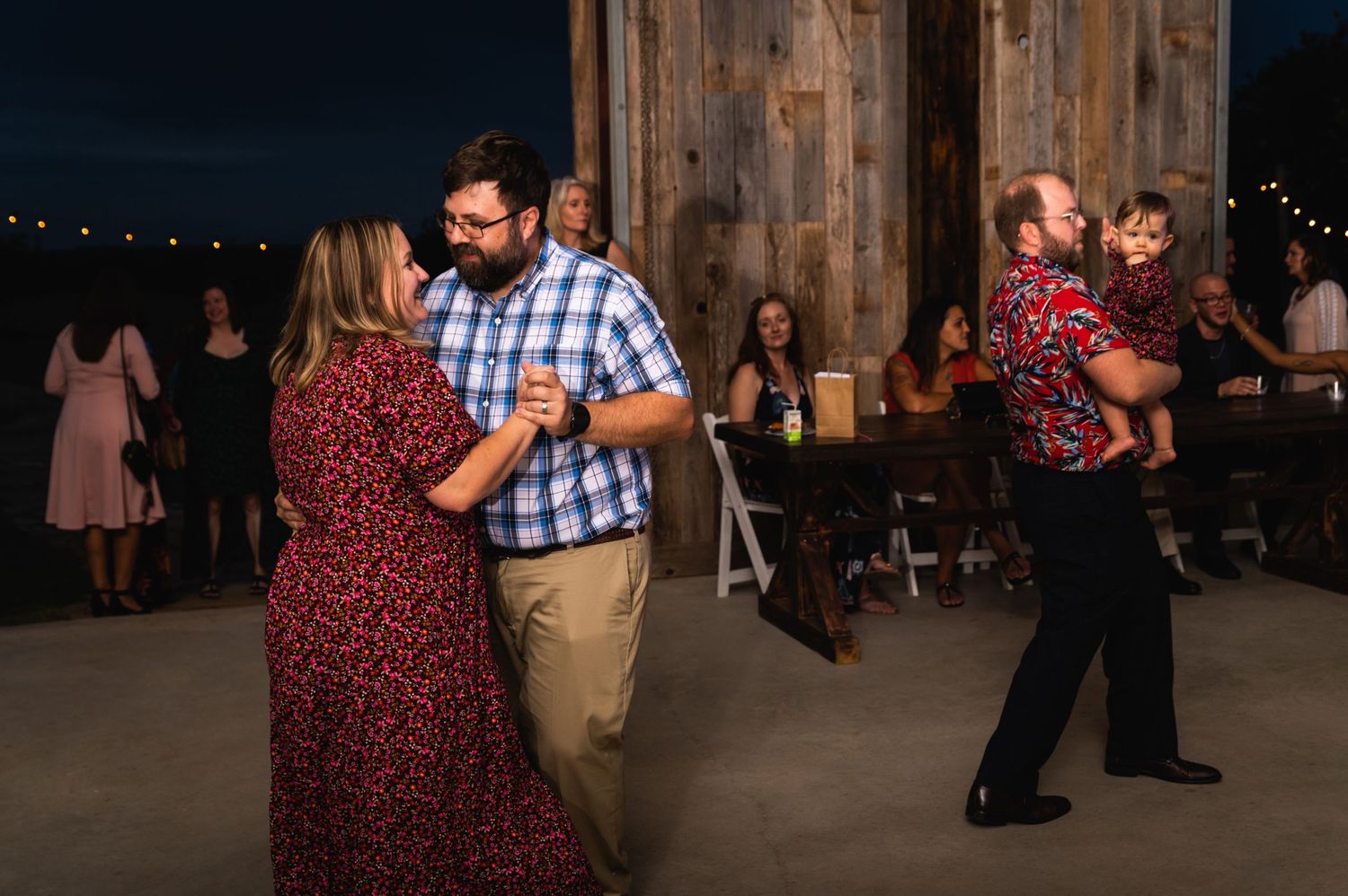 Guests dance together at a rustic barn wedding reception with wooden walls and string lights.
