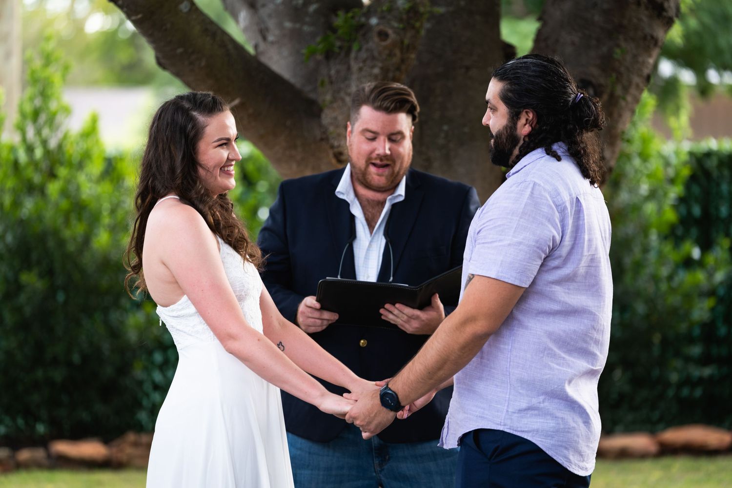 Outdoor wedding ceremony with officiant reading from clipboard under tree.